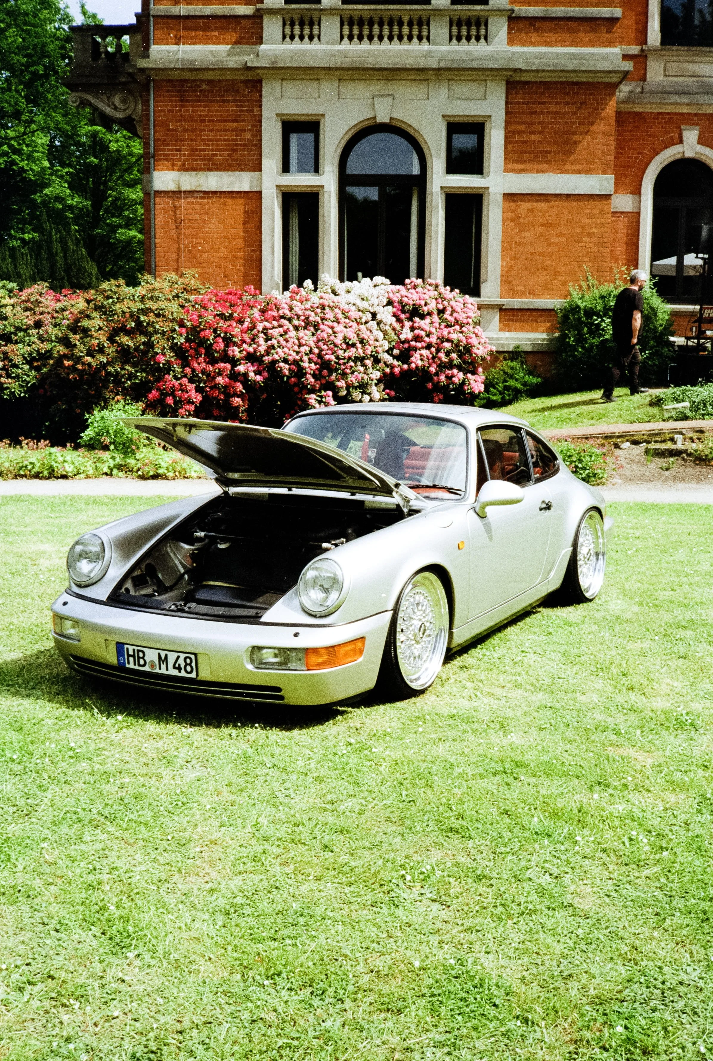 Silberner Porsche 911 mit offenem Motorhaube auf einem grünen Rasen vor einem großen roten Backsteingebäude mit vielen bunten Blumen und einer Person im Hintergrund.