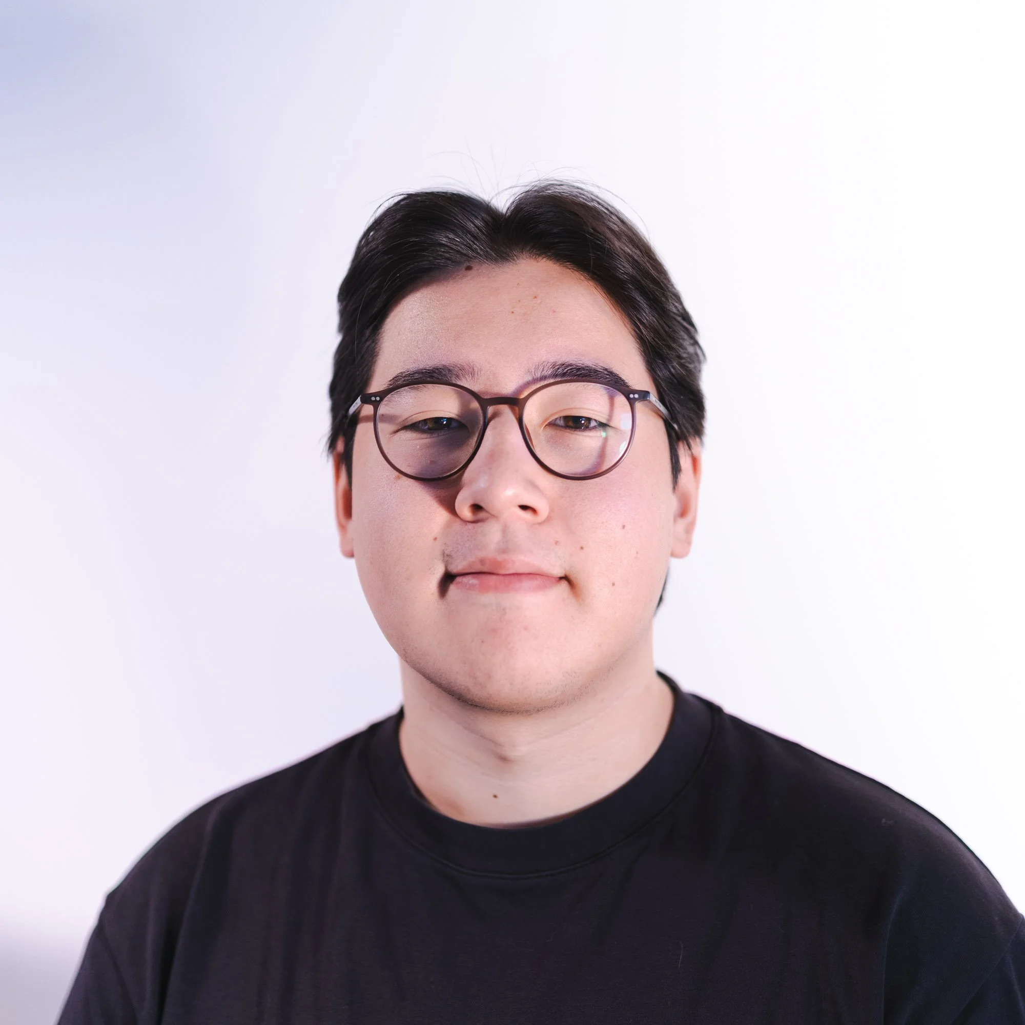 Porträt eines jungen Mannes mit schwarzen Haaren, Brille und schwarzem T-Shirt vor hellem Hintergrund.