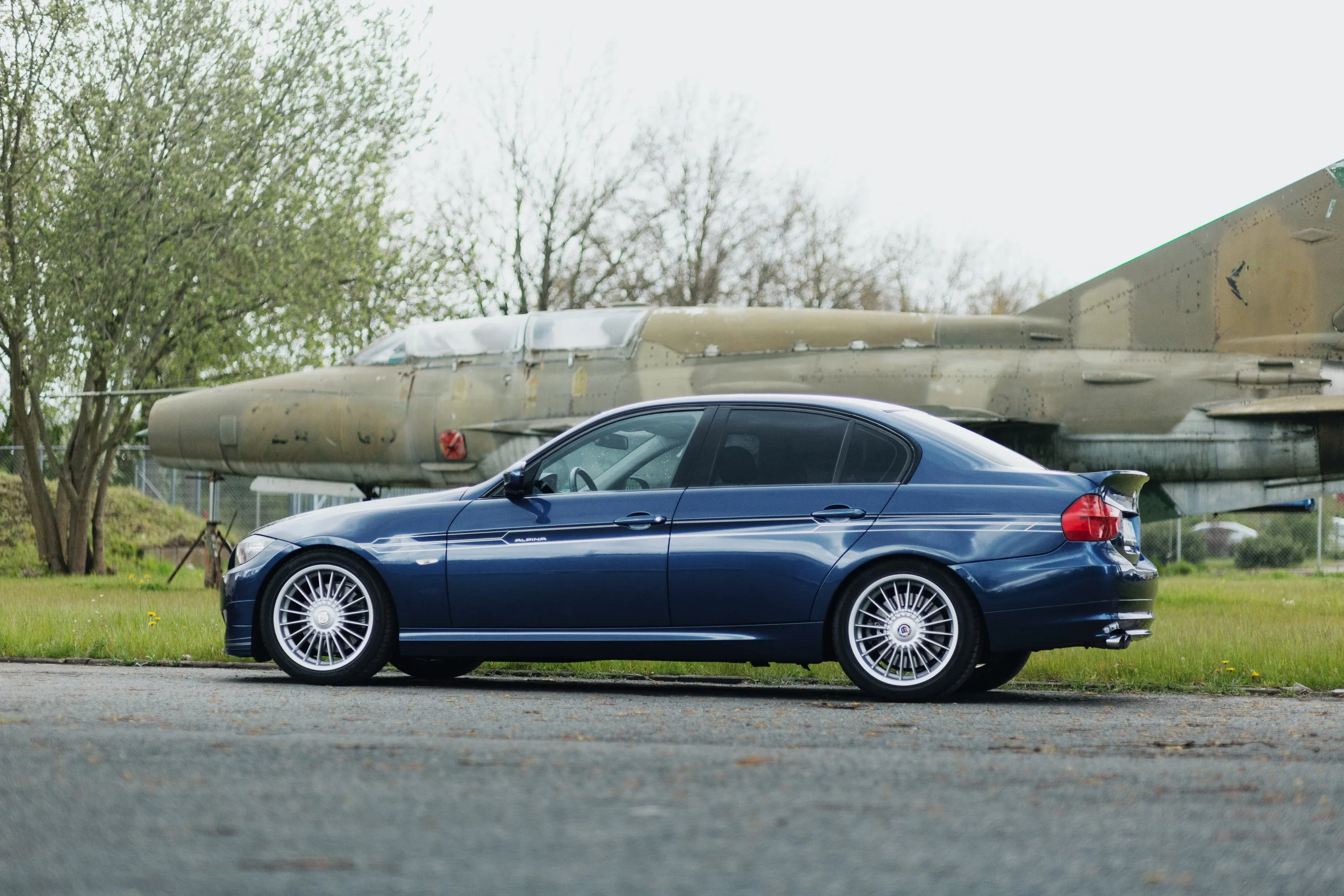Ein blauer BMW E92 3 Series Coupé steht auf einer Asphaltstraße vor einem camouflagefarbenen Kampfjet auf einer Wiese.