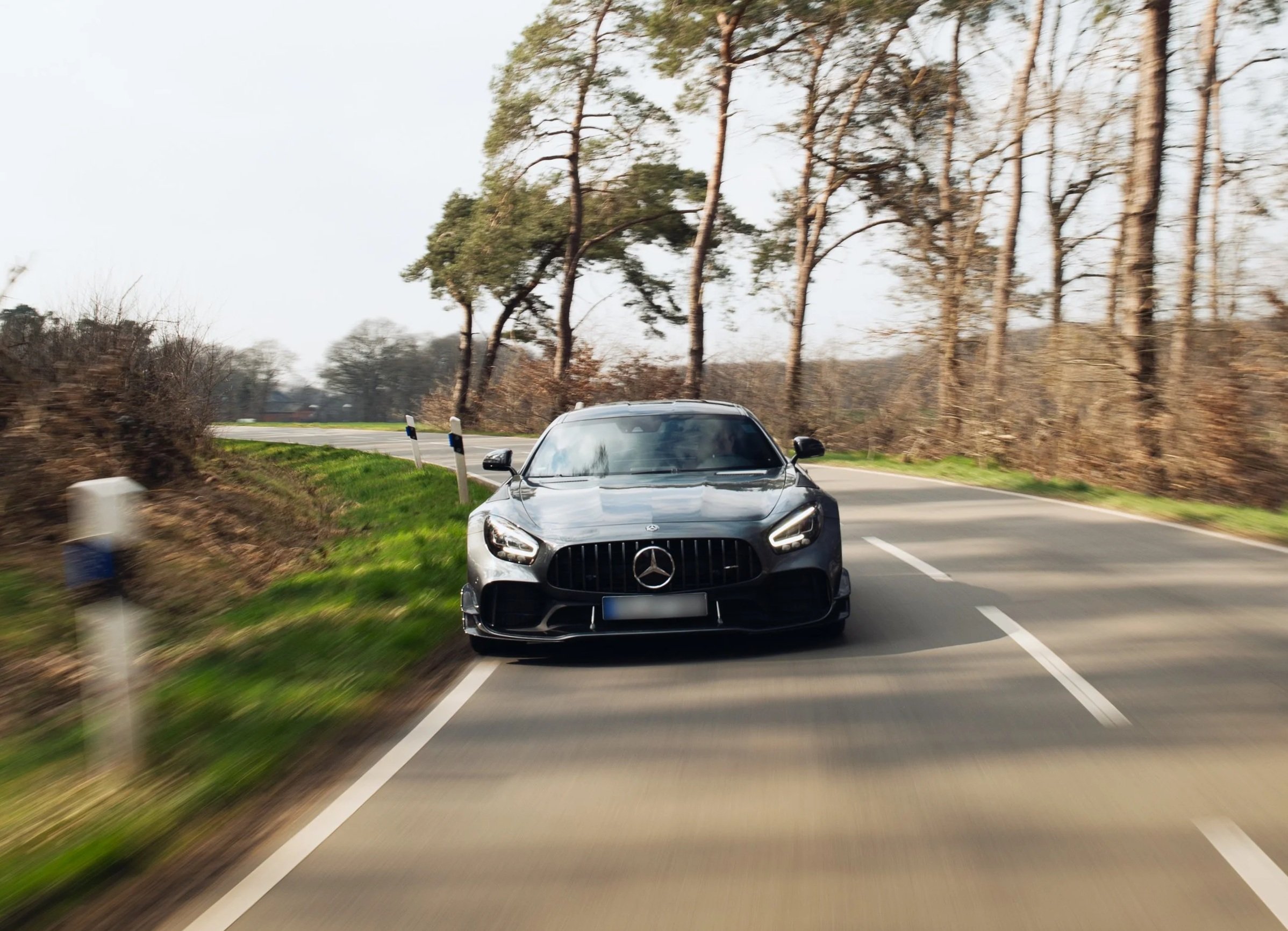 Ein schwarzer Mercedes-Benz Sportwagen fährt auf einer Landstraße zwischen Bäumen, mit grüner Wiese auf der linken Seite, bei Tageslicht.