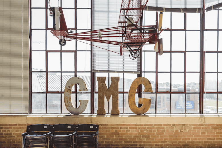 A vintage red airplane model suspended from the ceiling in front of large windows, with oversized letters 'C M G' placed on a brick ledge below, and a row of black chairs in front.