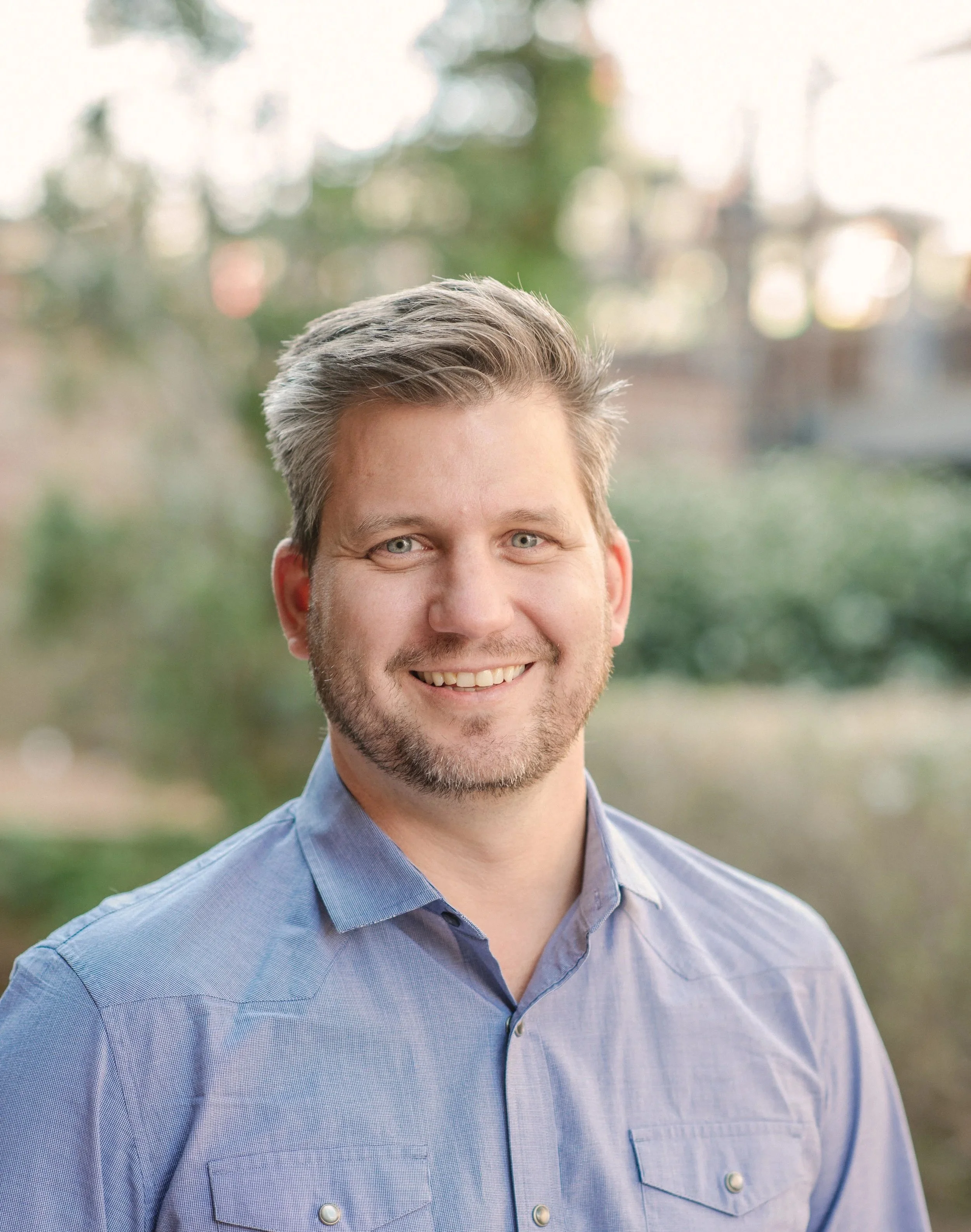 A man with gray hair and a beard smiling while wearing a light blue button-up shirt