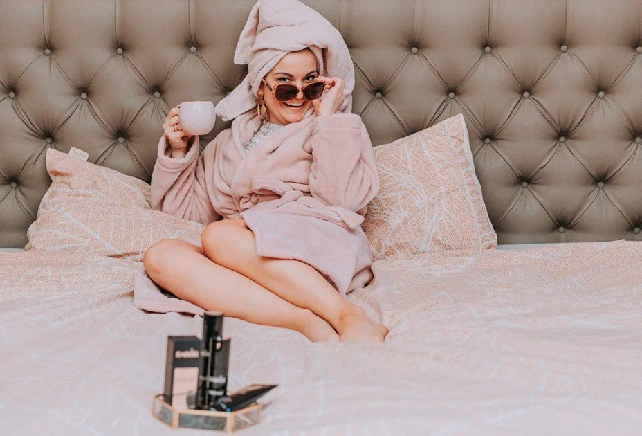 Vrouw in badjas met handdoek op hoofd, zittend op een bed met roze beddengoed, draagt zonnebril en houdt een kopje in de hand.