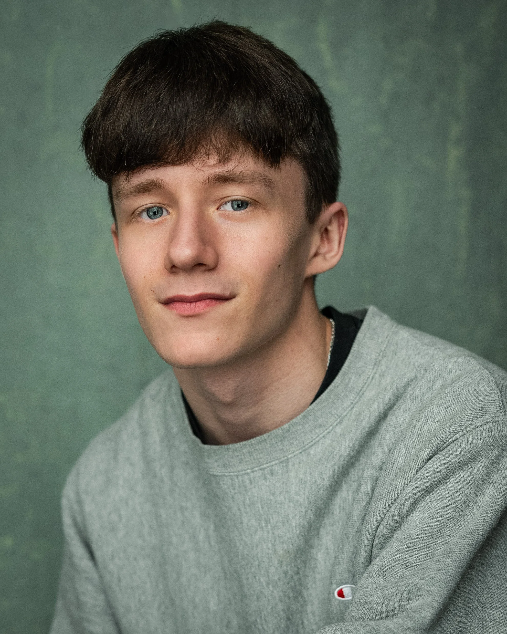 Portrait of a young man with brown hair and blue eyes wearing a gray sweatshirt against a green background.