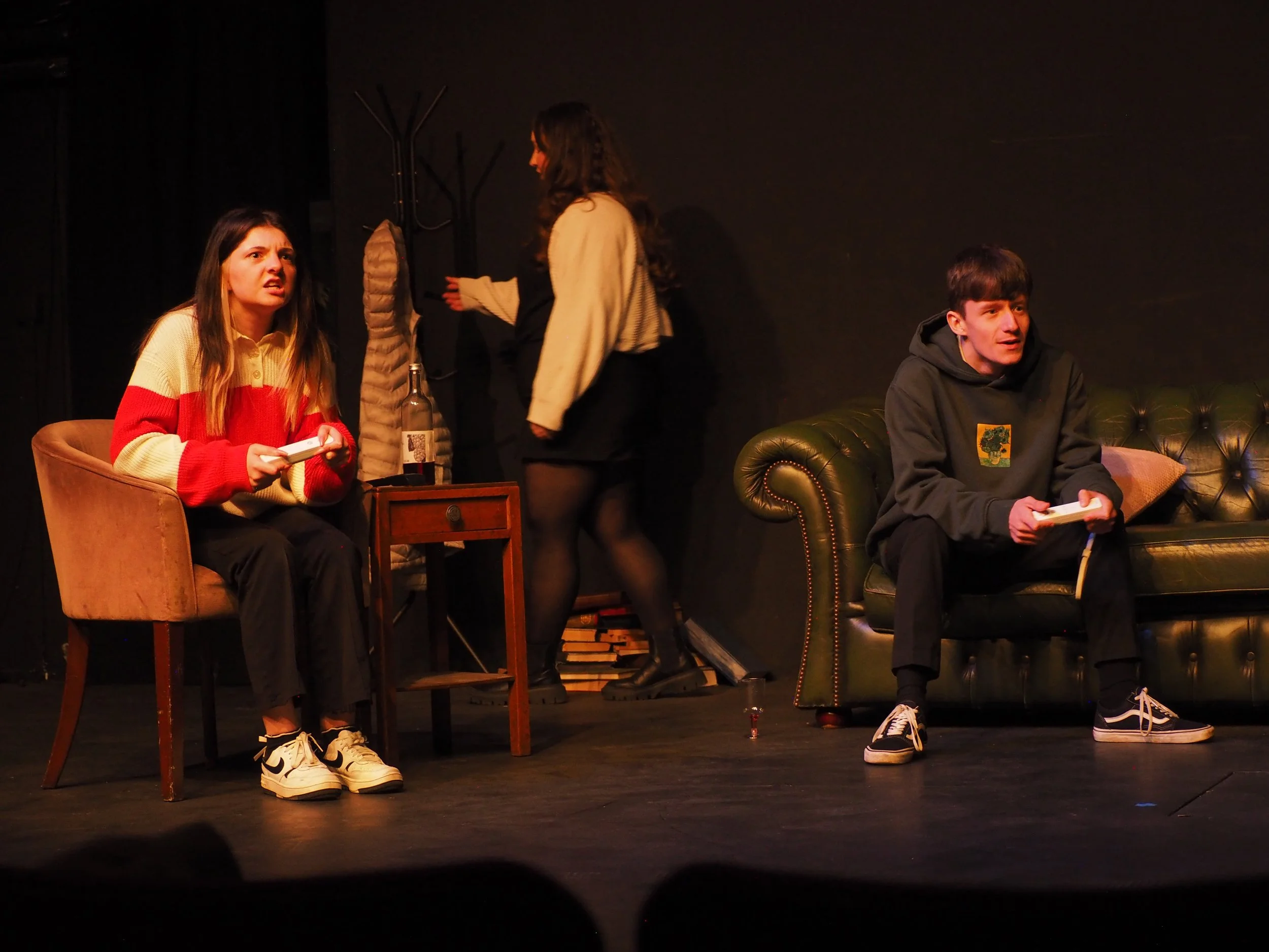 Three young actors on stage during a theatrical performance, two sitting on chairs with books, one standing with her back to the camera, in a minimalistic setting.