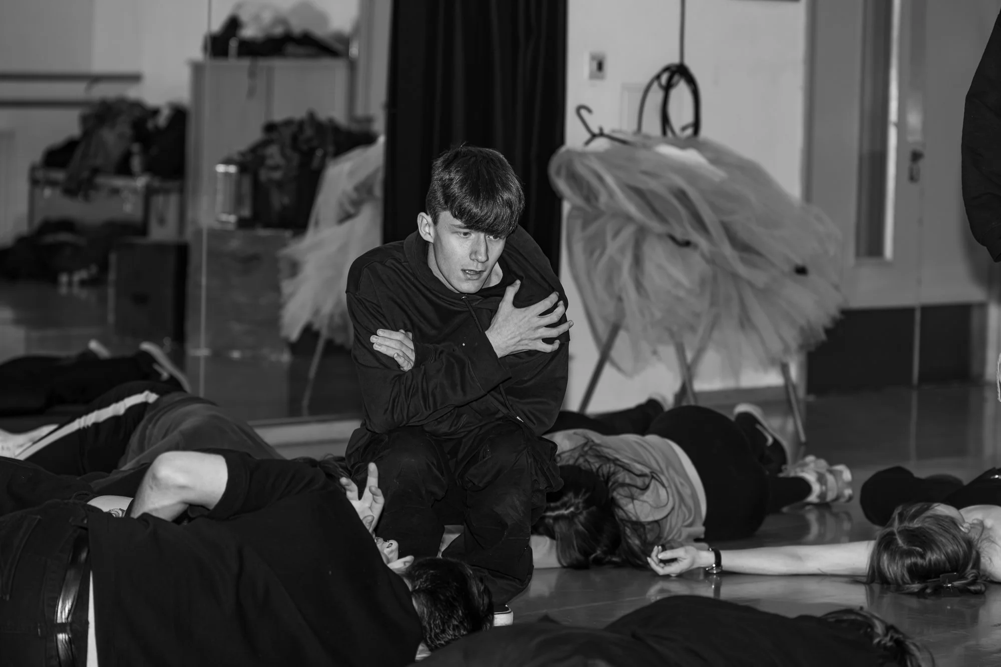 A young man appears shocked, kneeling on the floor while holding his chest, surrounded by people lying on the floor, suggesting a rehearsal in a drama studio.