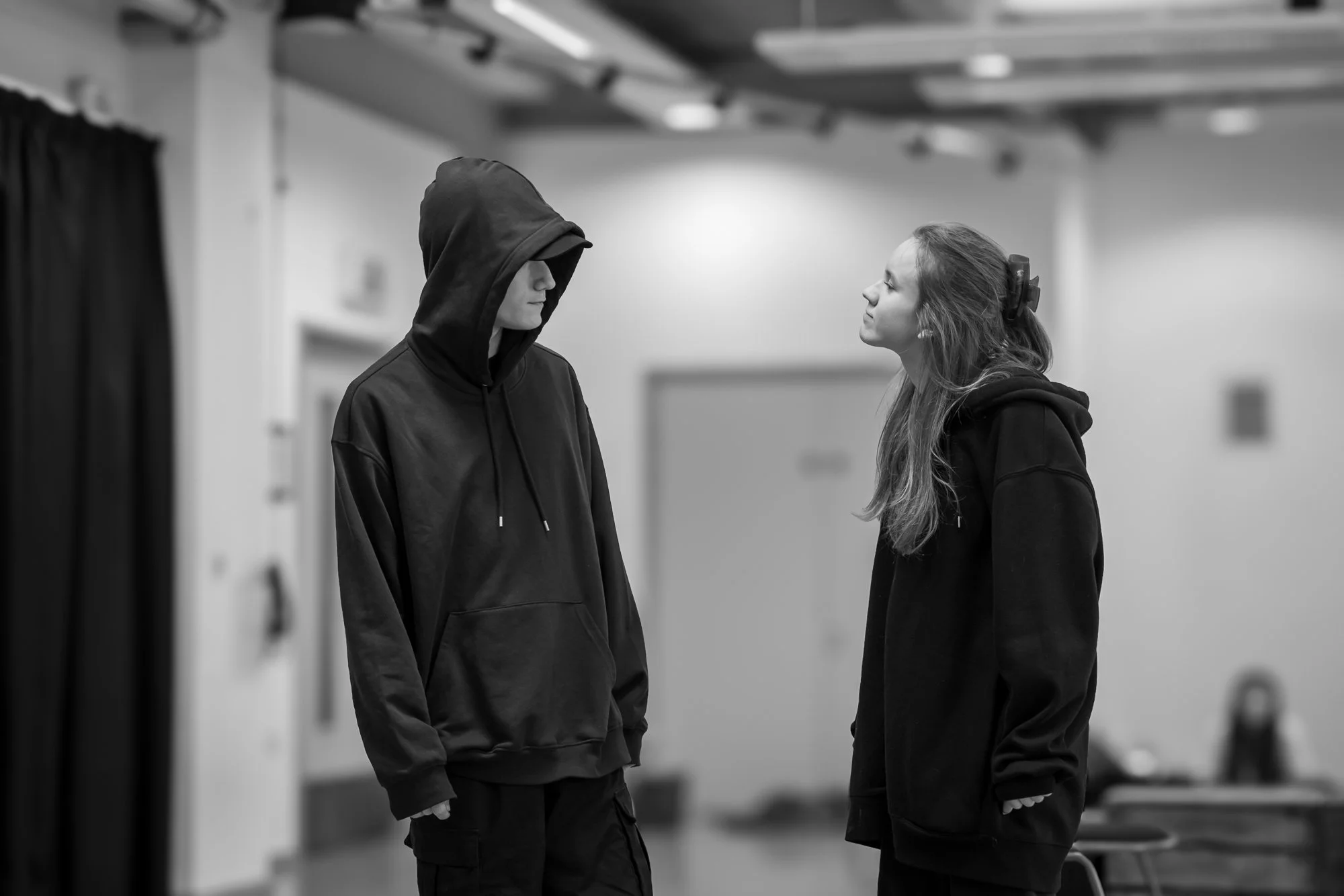 A young woman with long, wavy hair and a bow in her hair faces a person in a black hoodie with the hood up, indoors in a room with plain walls and some furniture, both appearing to be in a serious conversation.