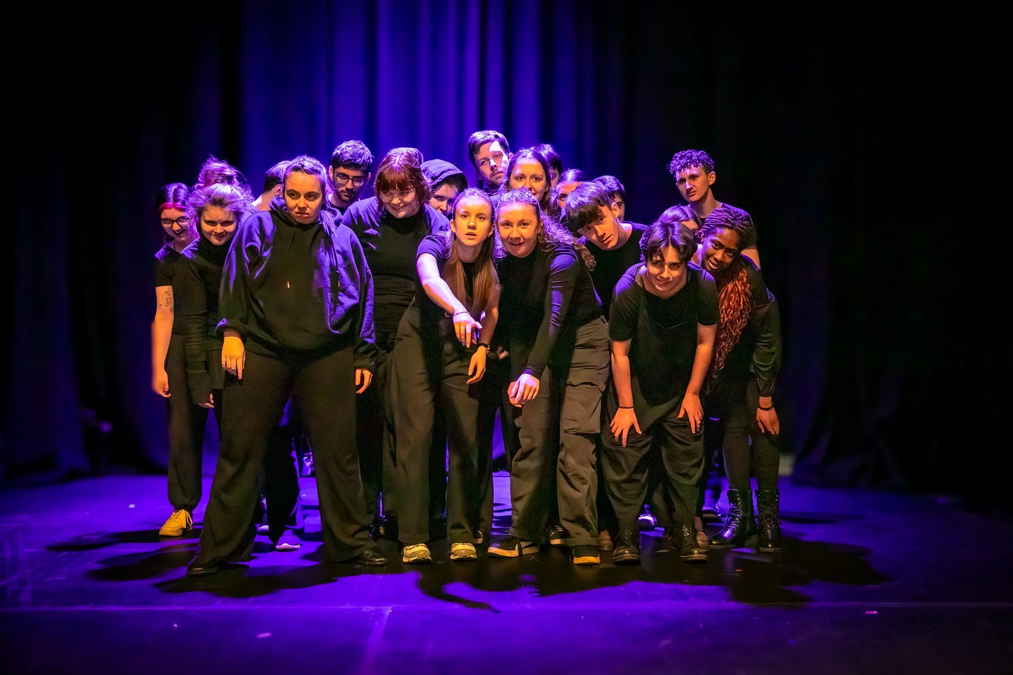 A group of young people on stage under purple and blue lighting, wearing mostly black, some looking serious, others smiling, with a dark curtain backdrop.