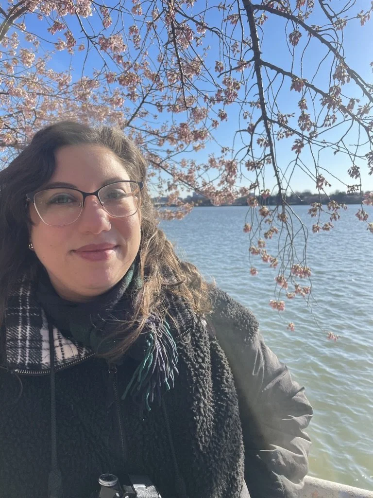 A woman with glasses and curly hair under a cherry blossom tree in front of the DC tidal basin water on a sunny day.
