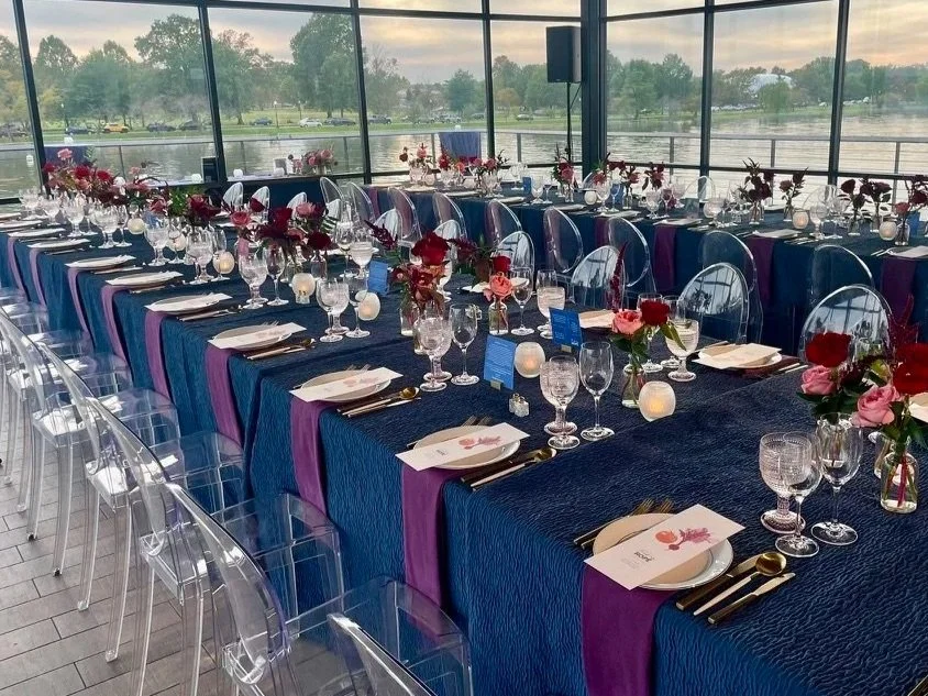 A banquet table decorated with dark blue and purple linens, with matching loral centerpieces and place settings, set in a glass-enclosed room overlooking a body of water.