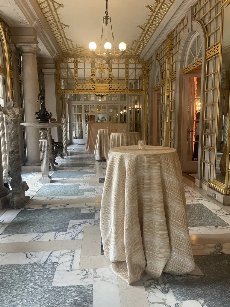 Elegant room with marble floor, gold accents, and tall windows. Several covered tables with lux beige tablecloths are arranged in the space. A chandelier hangs from the ceiling, and there is decorative architecture and sculptures along the walls.