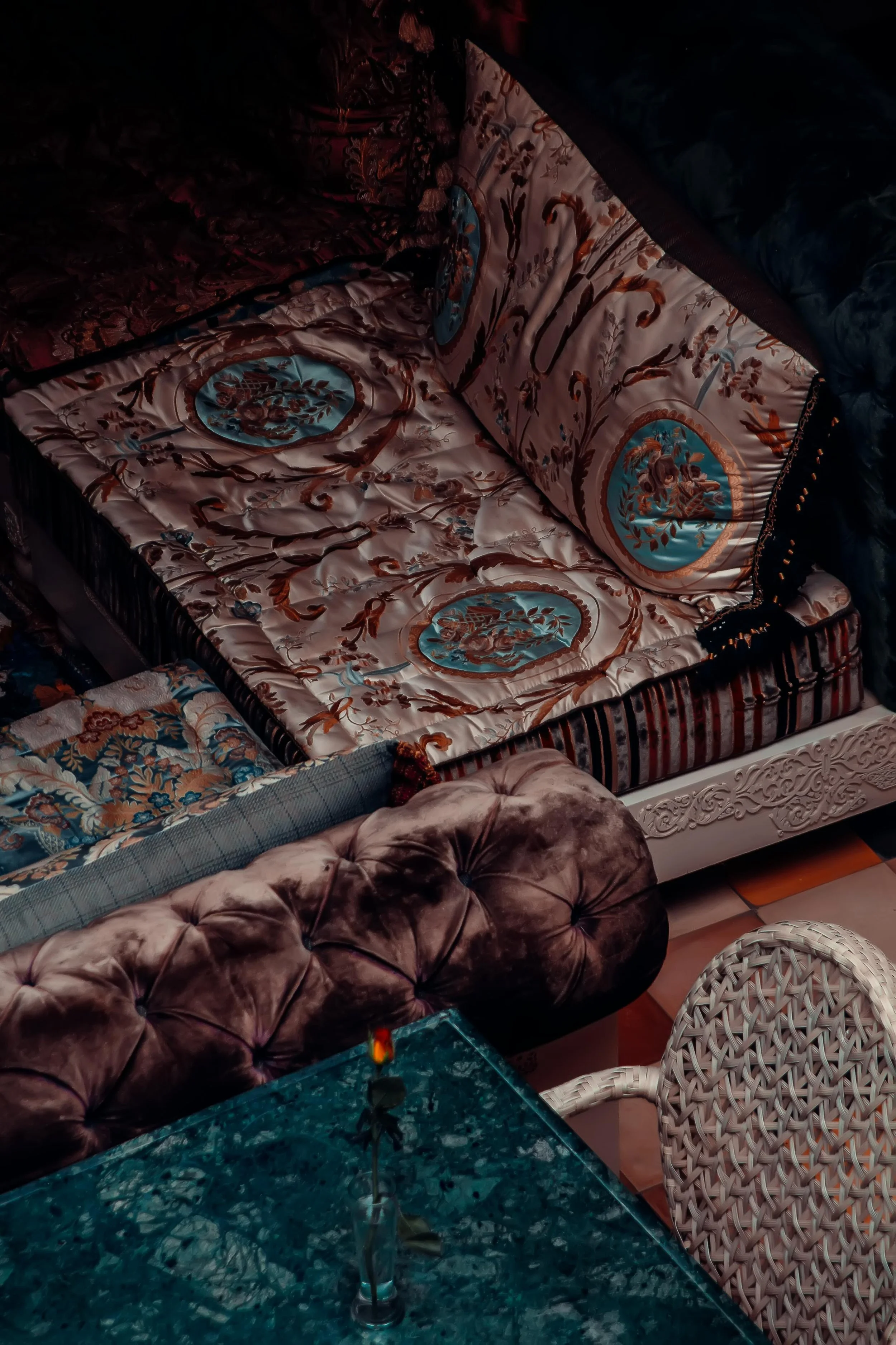 A vintage-style room with ornate patterned bedding on a bed, a tufted velvet armchair, a white woven chair, and a glass-topped table with a small flower in a vase.