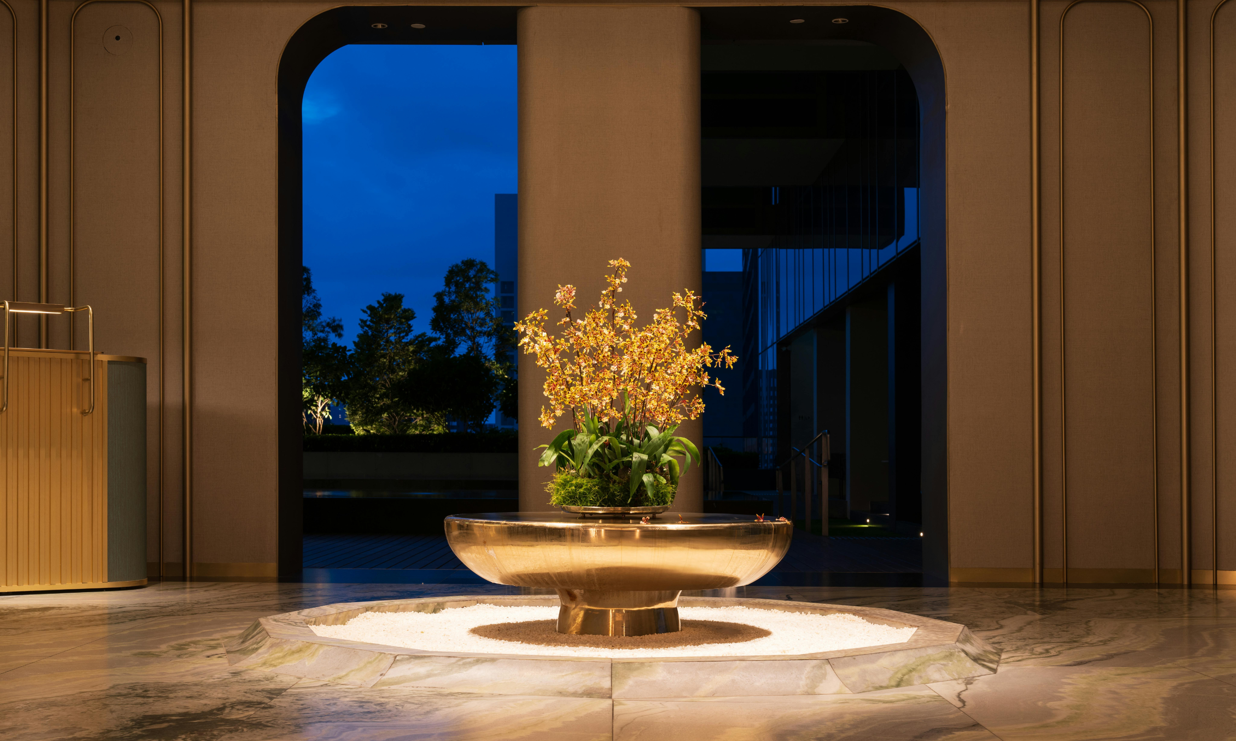 A decorative indoor floral arrangement with orchids in a copper bowl, illuminated with warm lighting, against a backdrop of an evening view through large arched windows showing a blue sky and trees.
