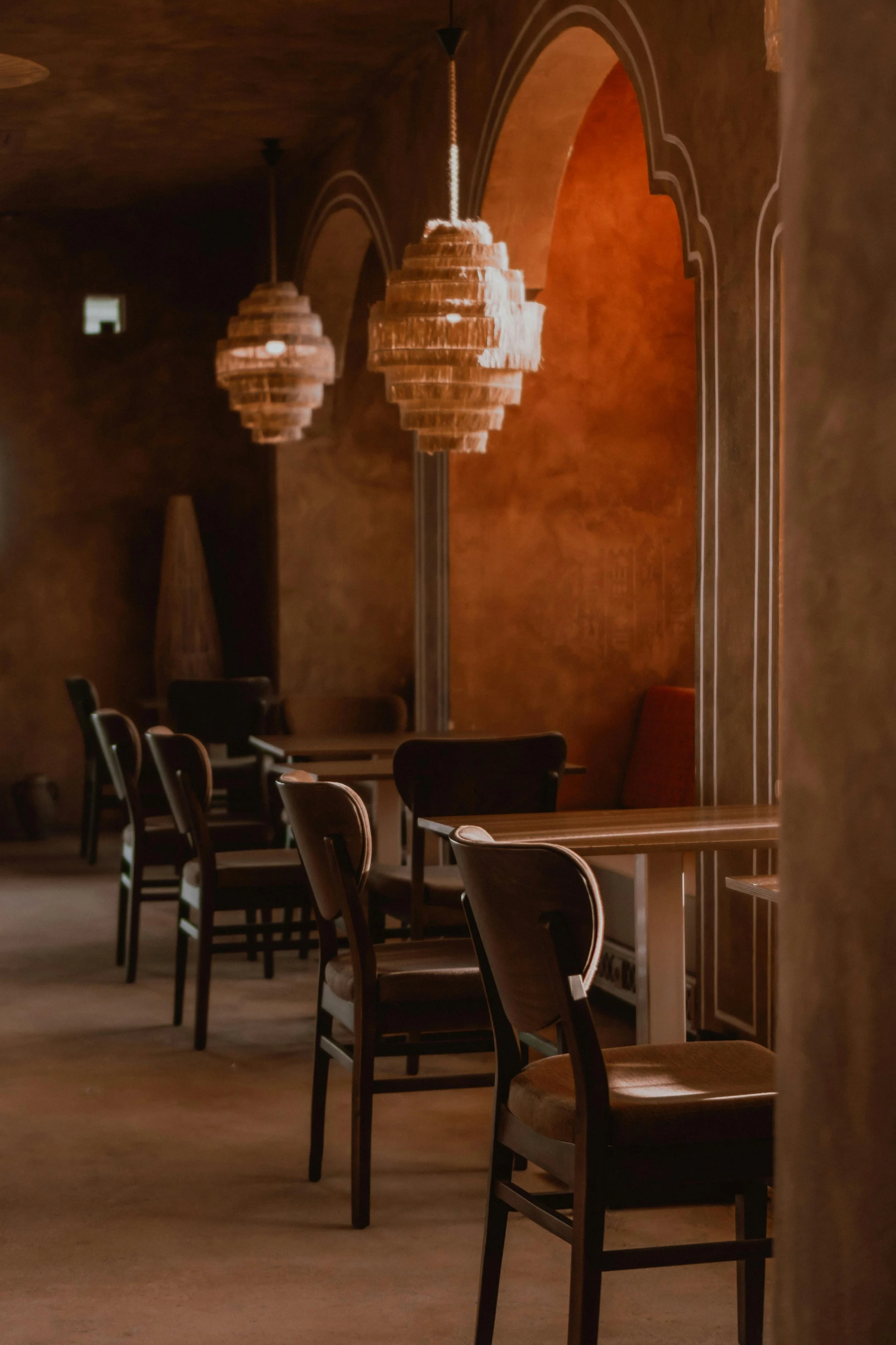 Dimly lit restaurant with wooden chairs, tables, and hanging chandeliers in a warm, earthy tone.
