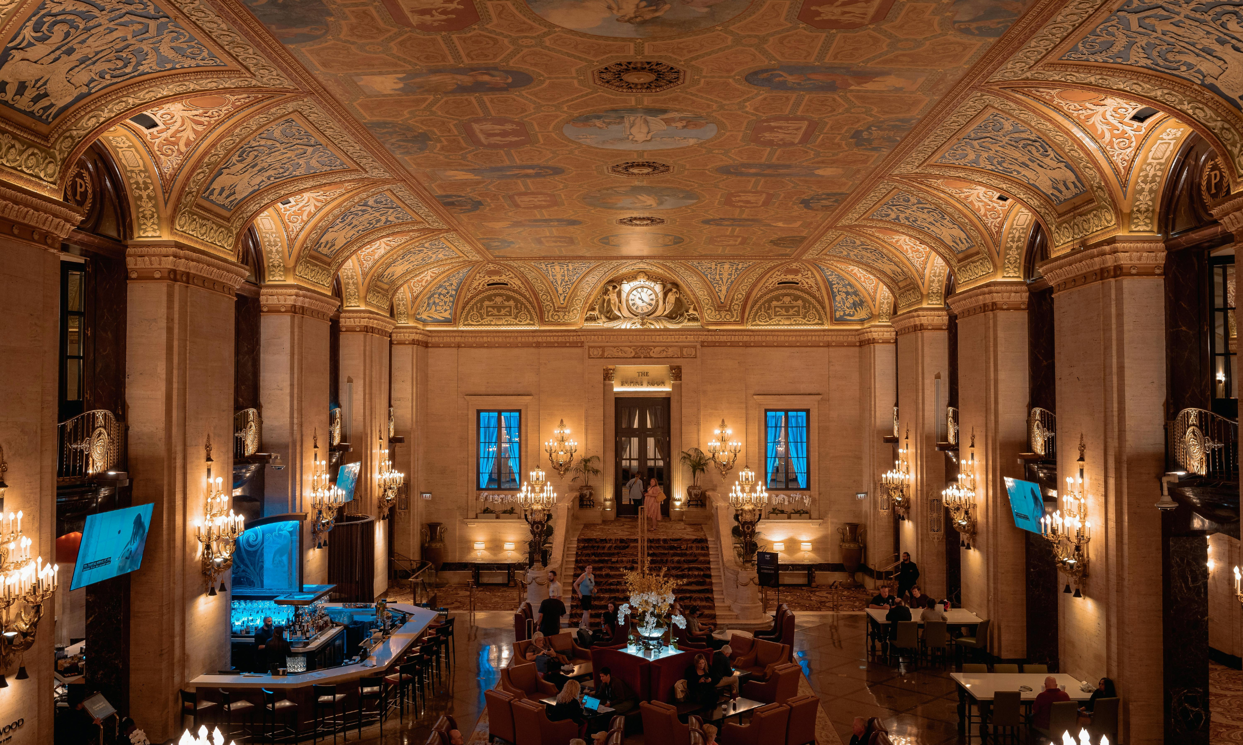 Elegant hotel lobby with high arched ceilings decorated with gold and blue patterns, chandeliers, and a central staircase. The area features seating with couches and tables, a bar with stools, and large windows with blue curtains.
