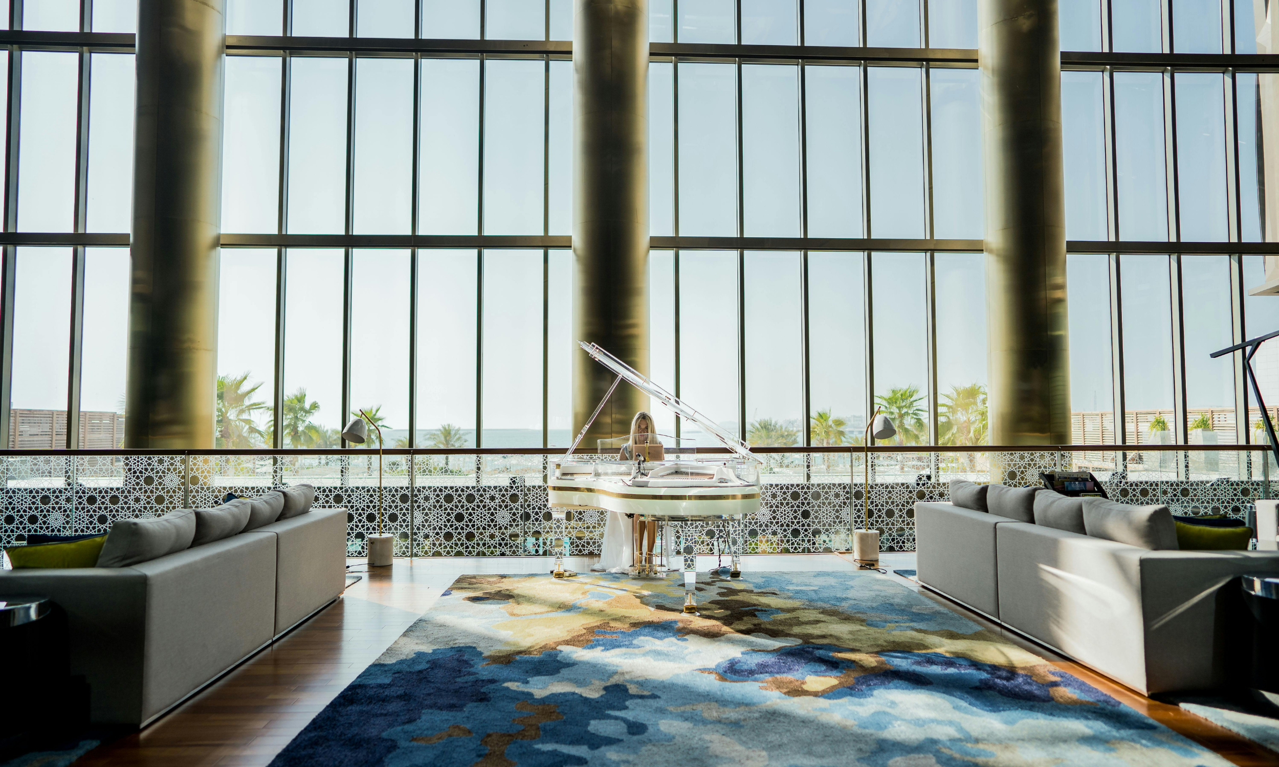 Interior of a spacious modern lounge with large floor-to-ceiling windows, a white grand piano, and comfortable gray sofas on a colorful area rug, overlooking a sunny outdoor area with palm trees.