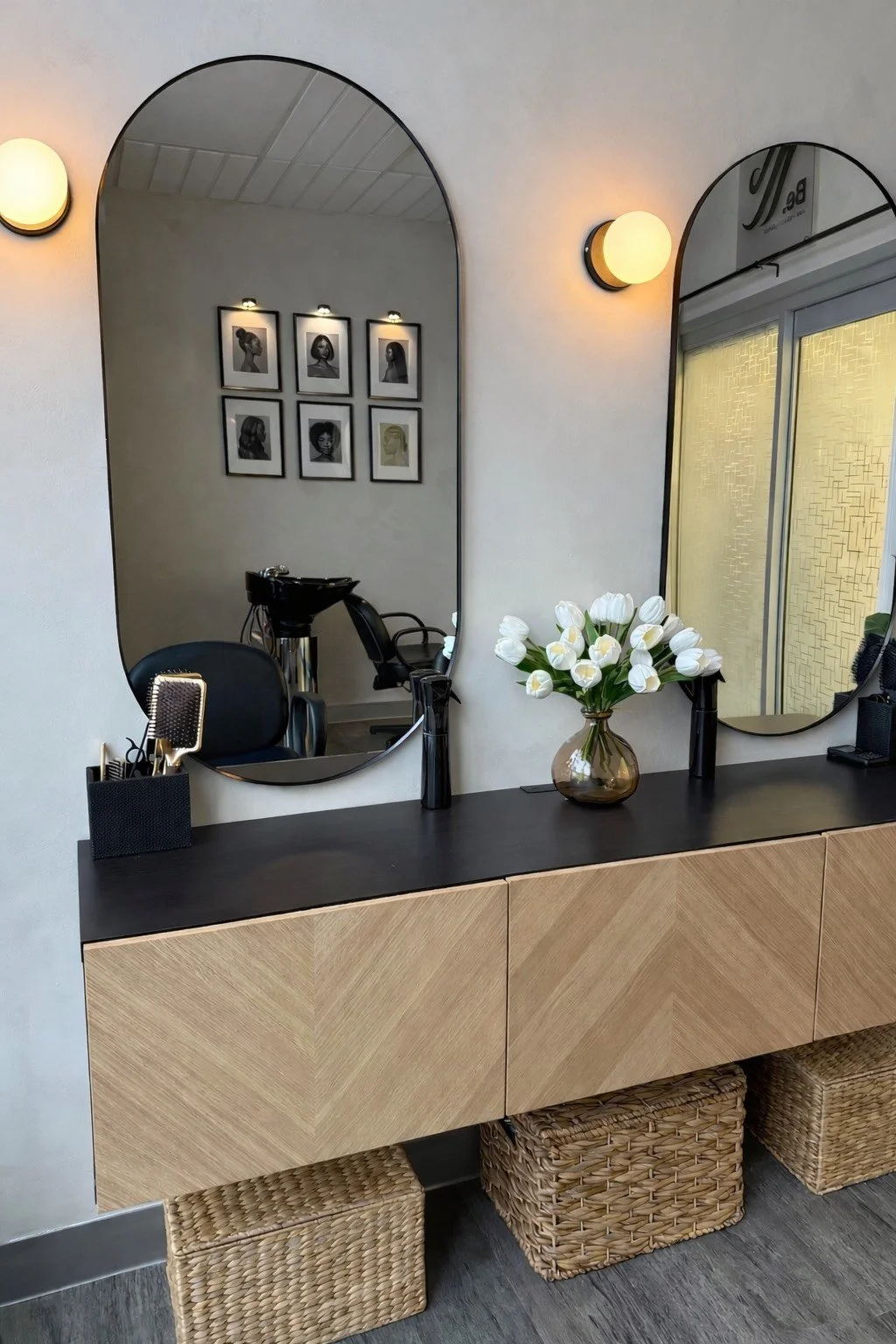 A salon station with two large oval mirrors, a black countertop, a vase of white tulips, a black hairbrush holder, and black styling products. Reflection in the mirrors shows framed black-and-white portraits of women, salon chairs, and a shampoo sink. Wicker baskets are beneath the cabinet, and wall-mounted round lights are on either side of the mirrors.