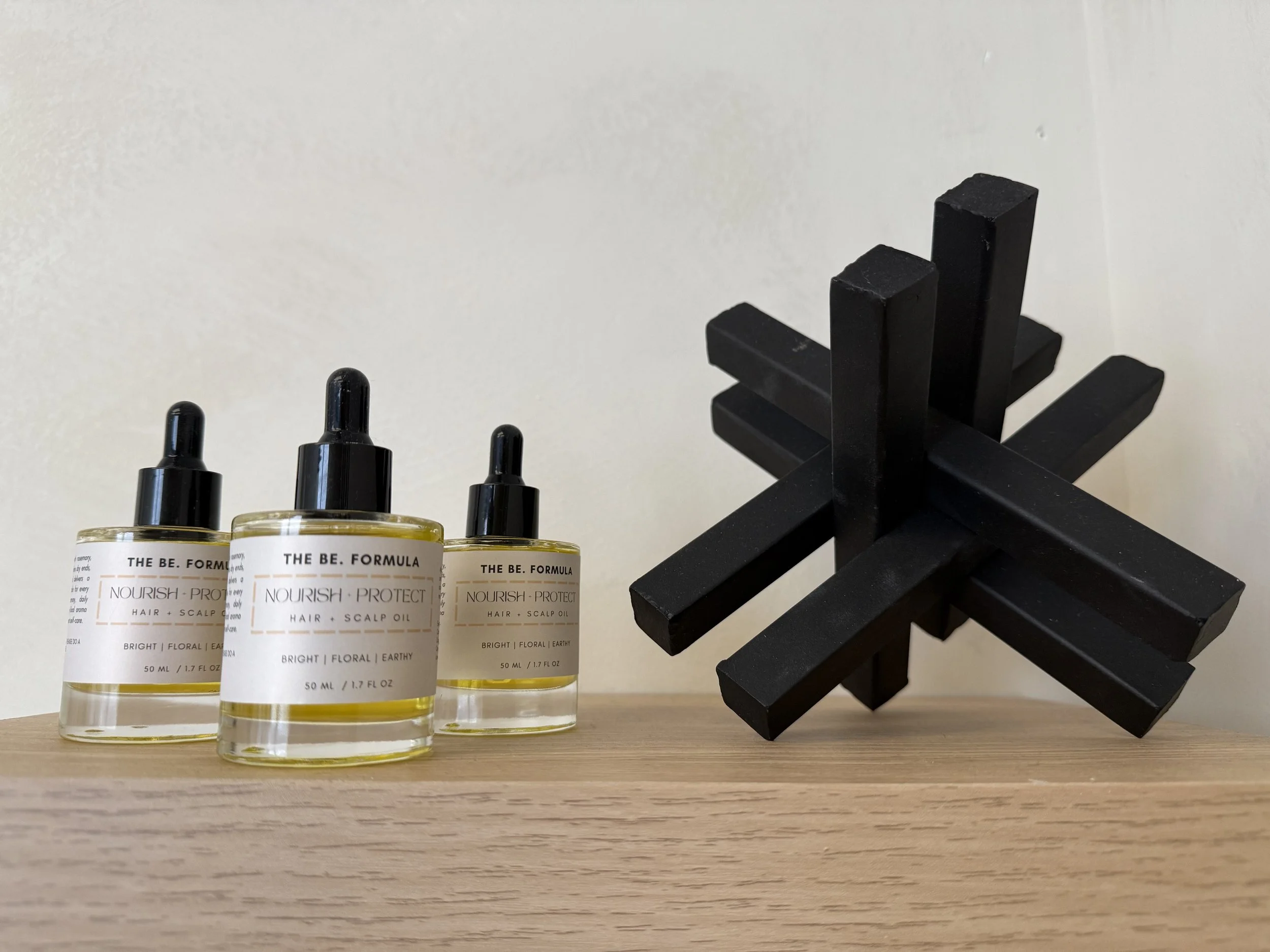 Three bottles of hair and scalp oil labeled 'Nourish Protect' placed on a wooden surface next to a black geometric decorative object.