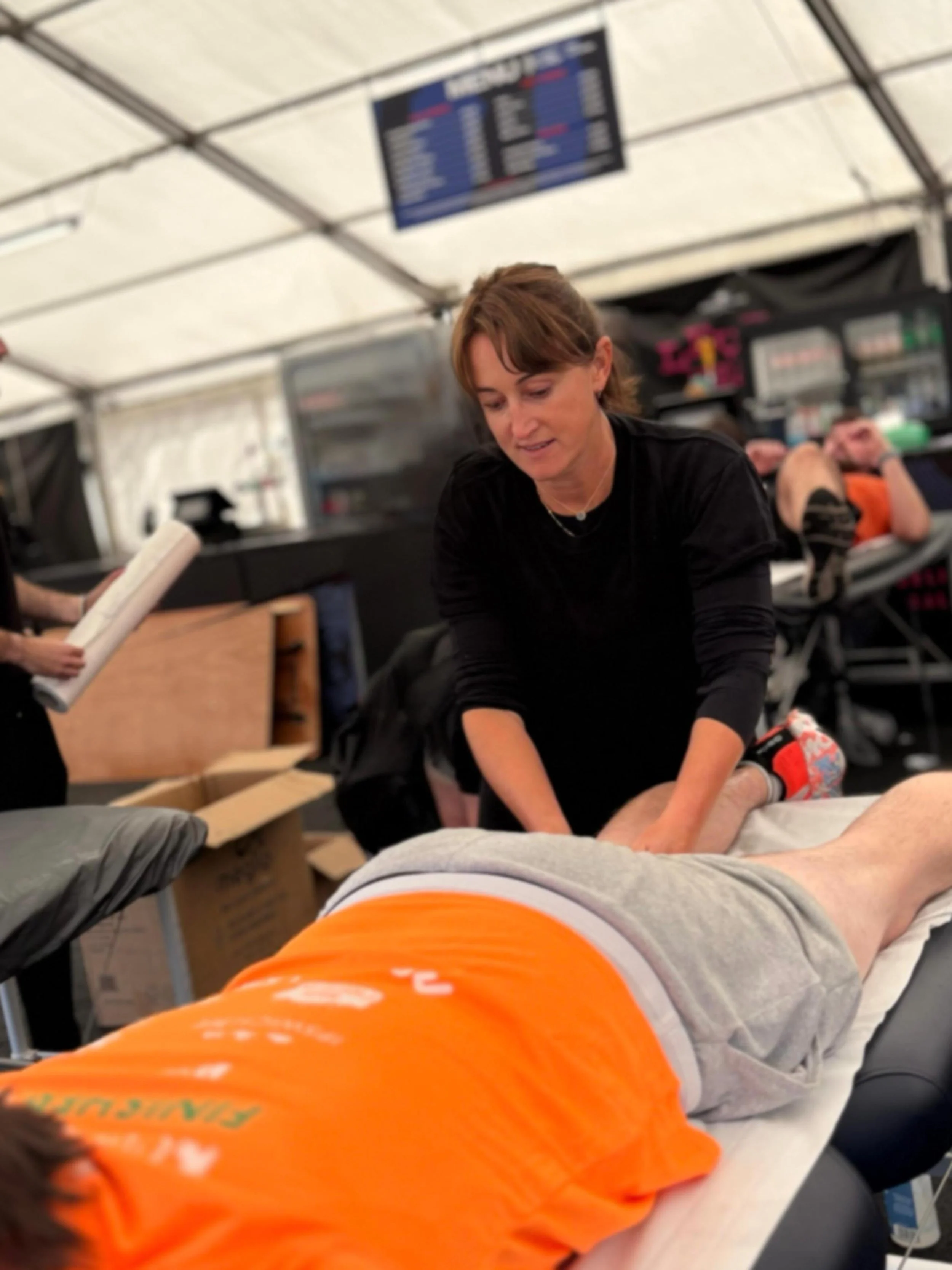 A woman providing massage therapy to a person lying face down on a massage table in a tent setting, with a digital departure board visible in the background.