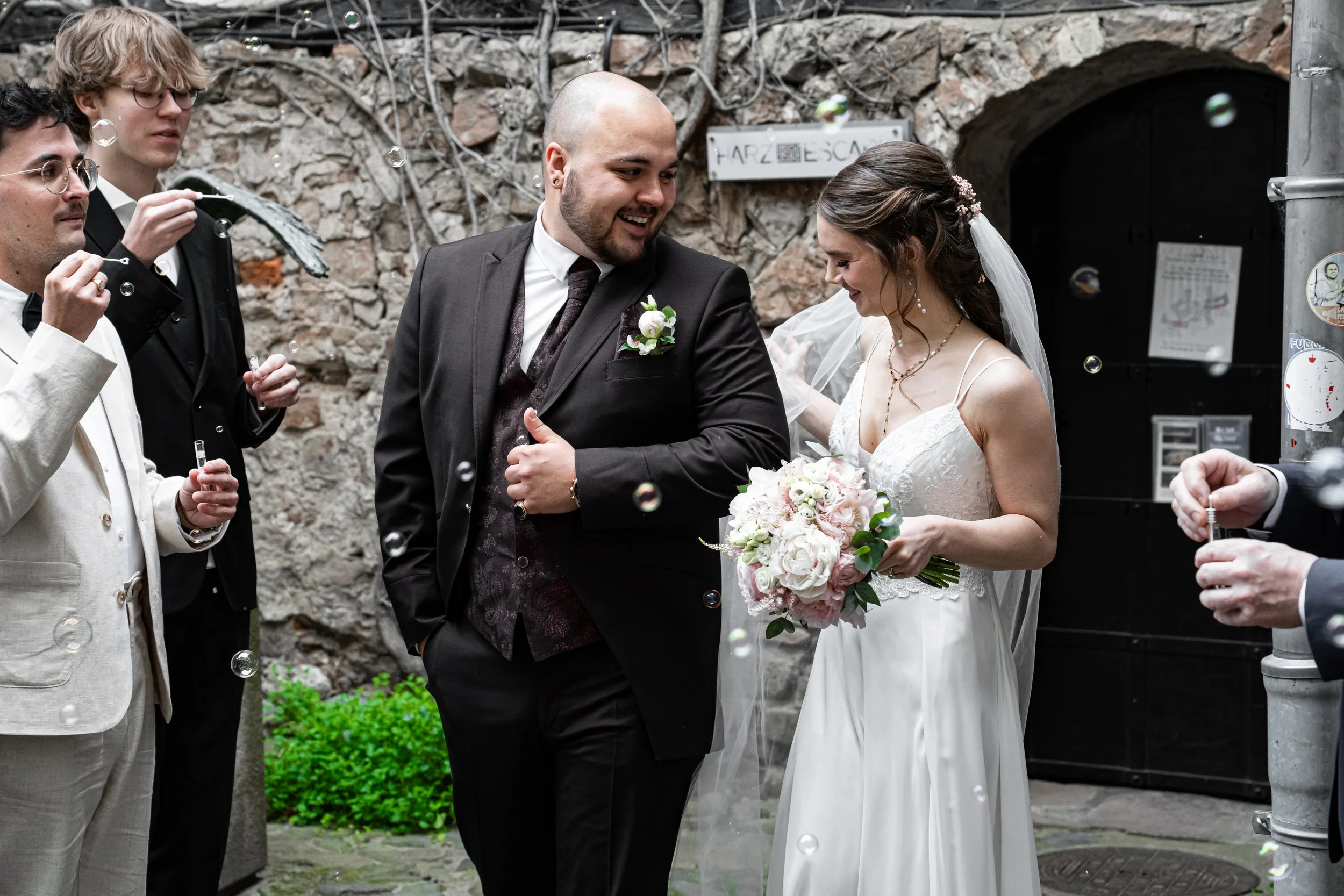 Ein Brautpaar steht bei ihrer Hochzeit, umgeben von Gästen, die Seifenblasen machen. Die Braut trägt ein weißes Hochzeitskleid mit Blumenstrauß, der Bräutigam trägt einen schwarzen Anzug. Die Szene findet vor einer Steinmauer und einem schwarzen Tor statt.