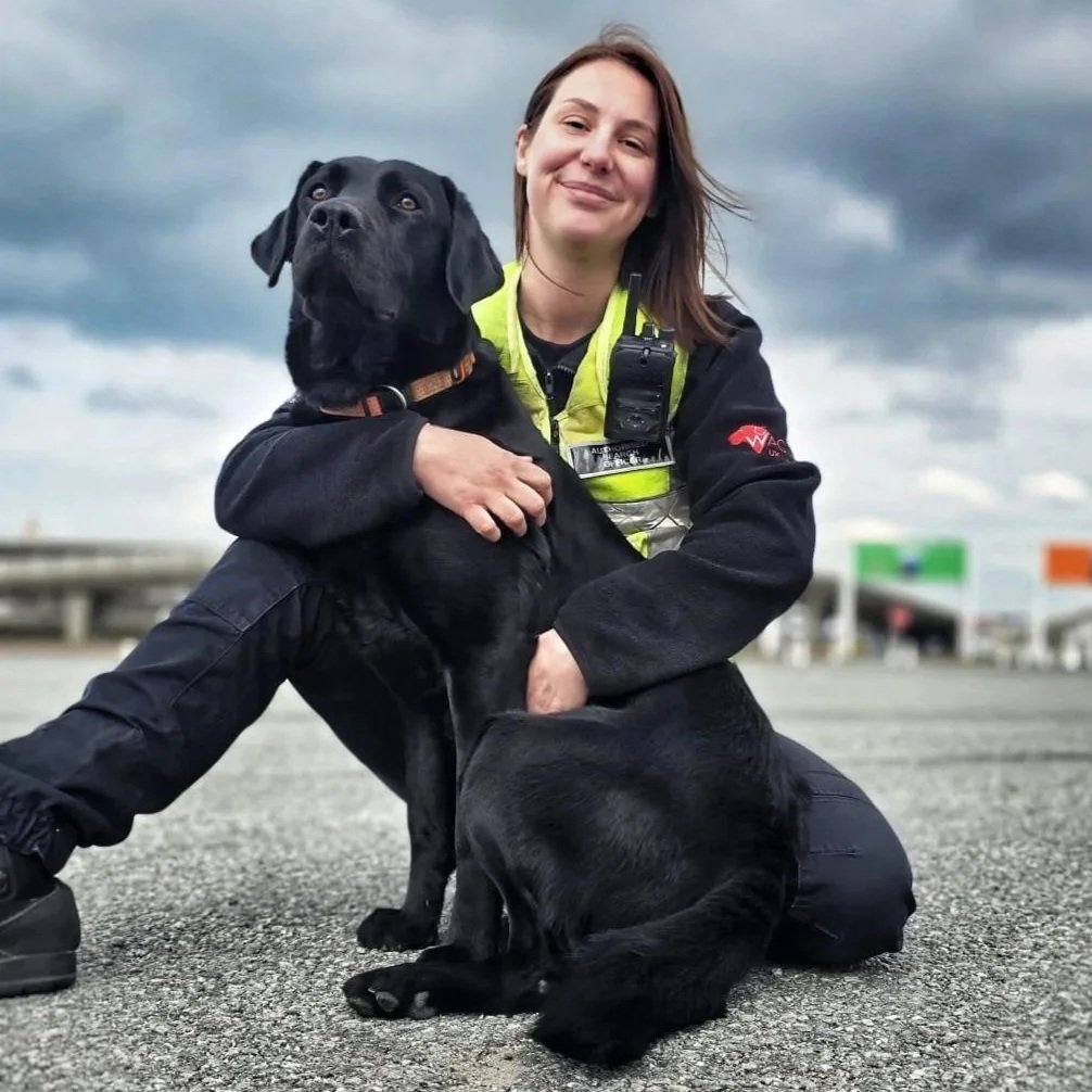 A woman and a black Labrador retriever dog sitting on pavement outdoors with cloudy sky in background. The woman is smiling and wearing a black jacket with yellow-green reflective safety vest and kneeling, holding the dog.