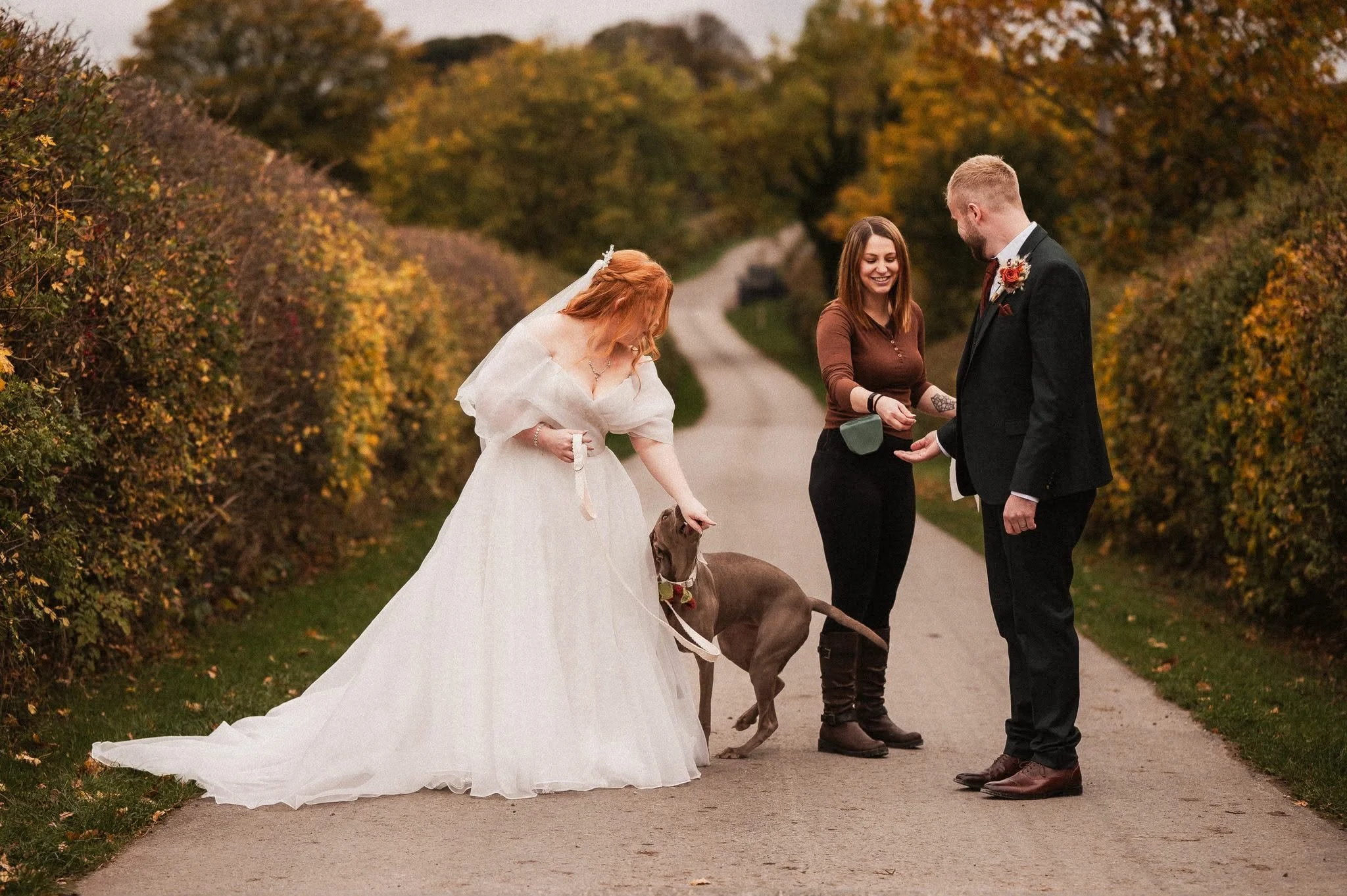 A wedding scene in an outdoor setting with a bride in a white gown, a groom in a black suit, and a woman standing between them, holding a dog on a leash. The woman appears to be performing a wedding ritual, and the background shows a gravel path lined with autumn-colored foliage.