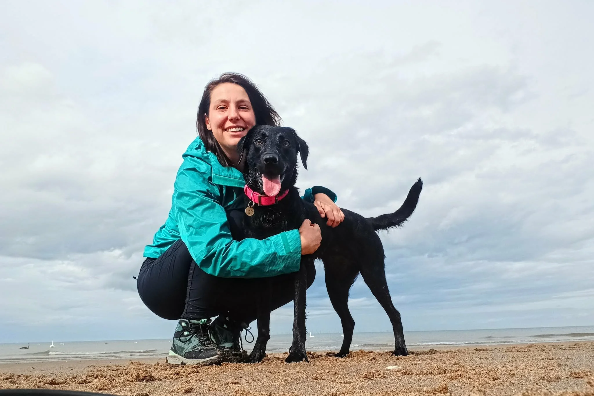 A woman smiling and hugging a black Labrador on a beach with overcast sky.