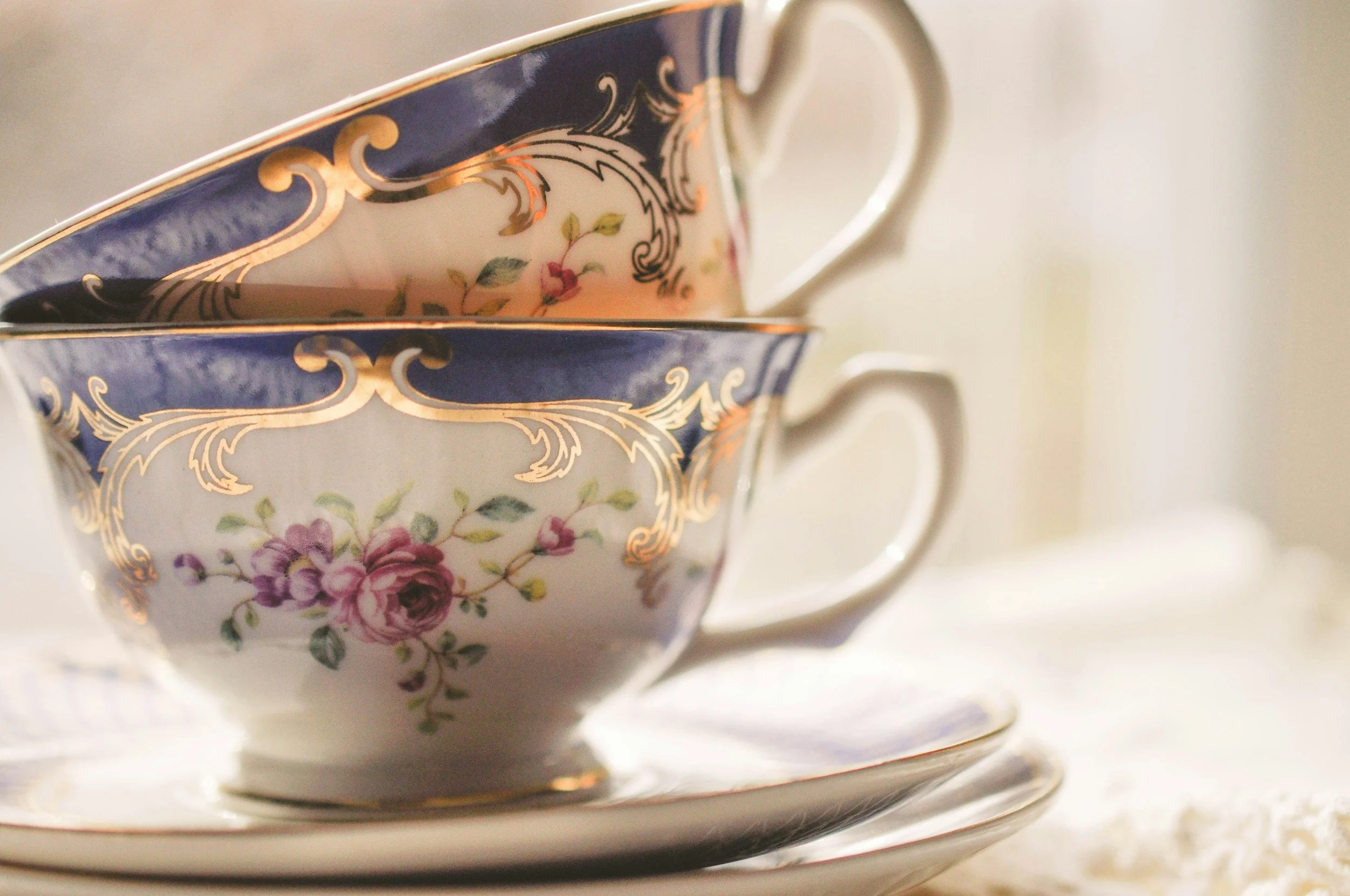 Close-up of a decorative porcelain teacup stacked on a saucer, featuring floral patterns with pink and purple flowers, gold accents and a dark blue border, with a plain background.