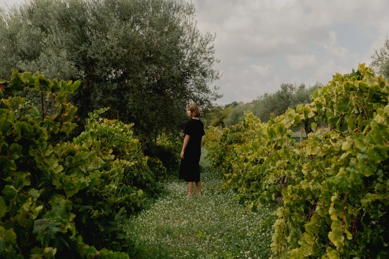 A woman in a black dress standing on a grassy path through a vineyard with lush green grapevines and trees under a cloudy sky.