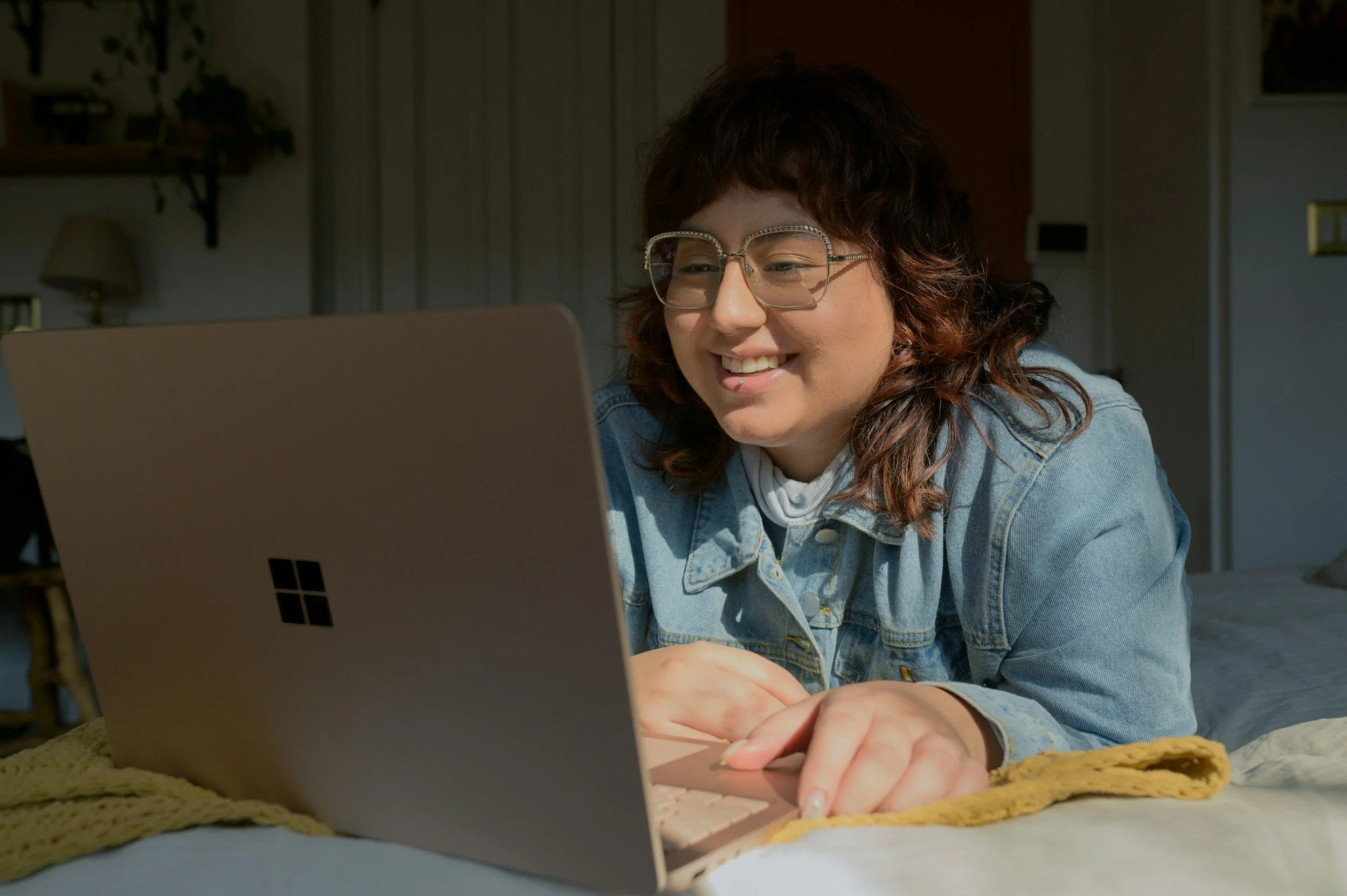 A young woman with glasses and a denim jacket smiling while using a laptop at a table.