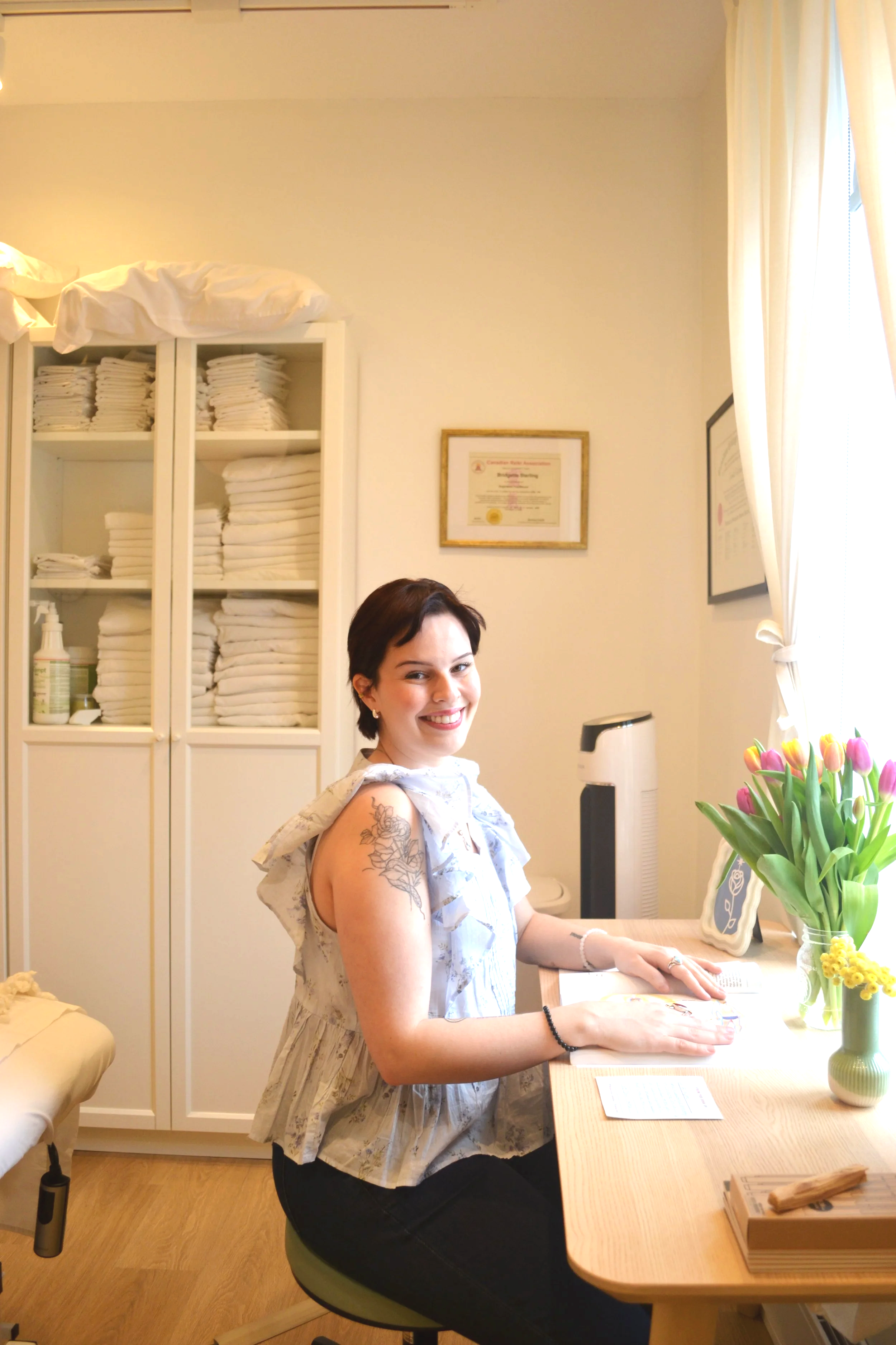 A woman with short dark hair, wearing a sleeveless dress with floral patterns, is sitting at a light wooden desk in a room with white cabinets full of folded towels. There is a framed certificate or diploma on the wall and a vase of colorful tulips on the desk. She is smiling and has tattoos on her upper arm.