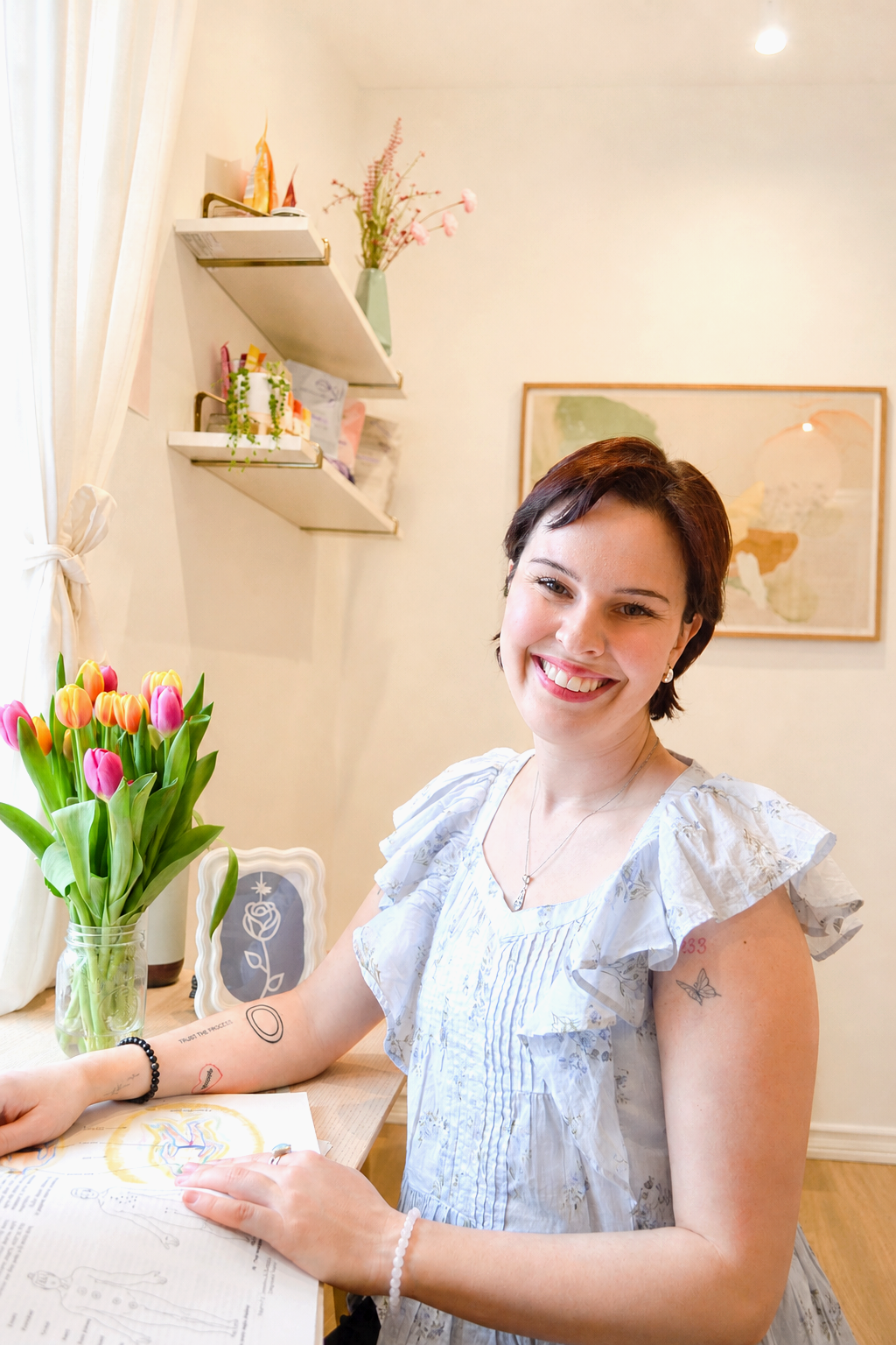 A woman with short brown hair smiling while sitting at a desk with a newspaper in front of her. She has colorful tattoos on her arms, including a butterfly on her right arm and a red design on her left arm. She wears a white ruffled blouse and jewelry. A vase of pink and yellow tulips is on the desk, and decorative shelves with plants and artwork are on the wall behind her.