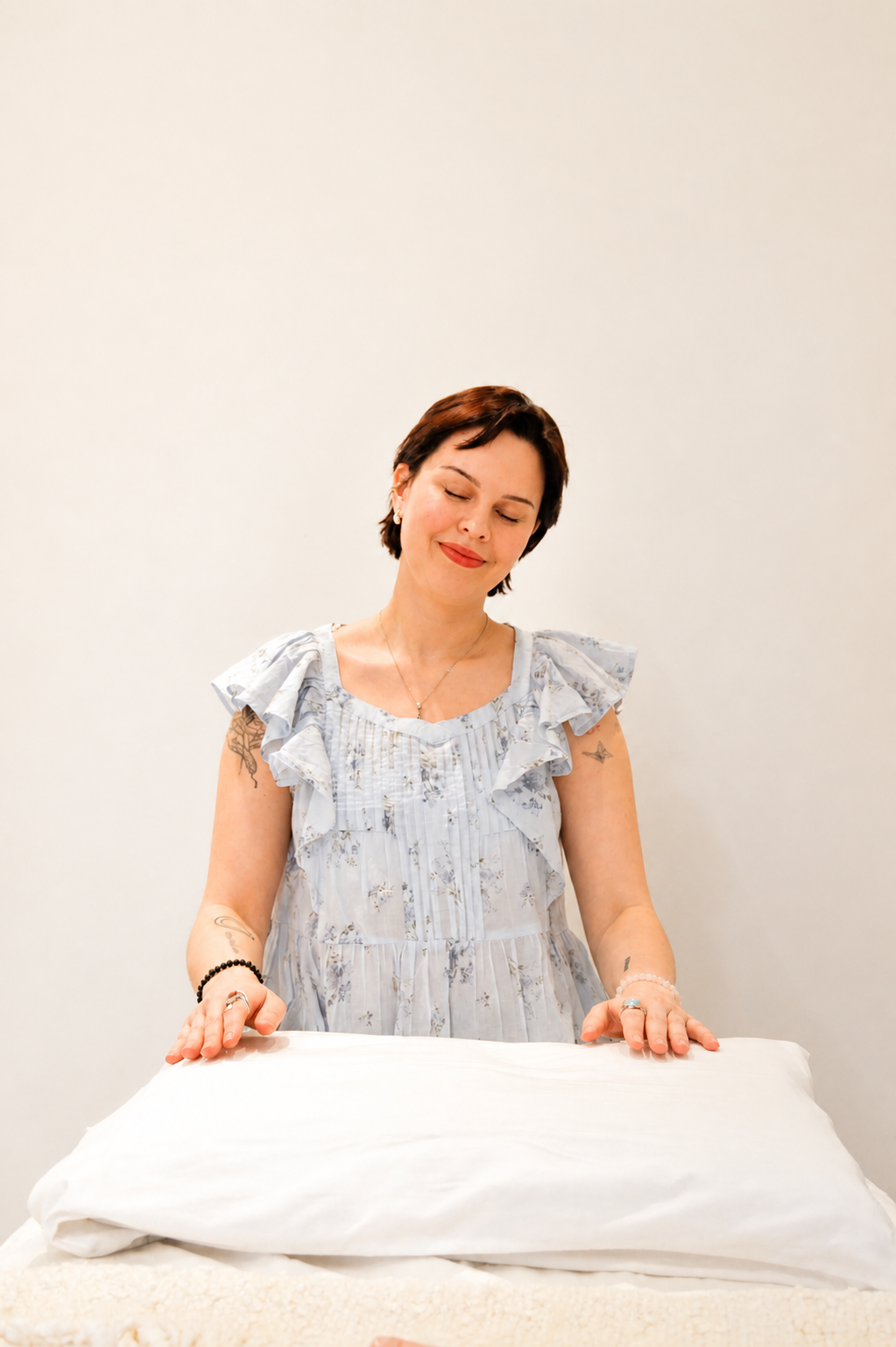 A woman with short brown hair and tattoos on her arms, smiling with her eyes closed, wrapping a bed with white sheets.
