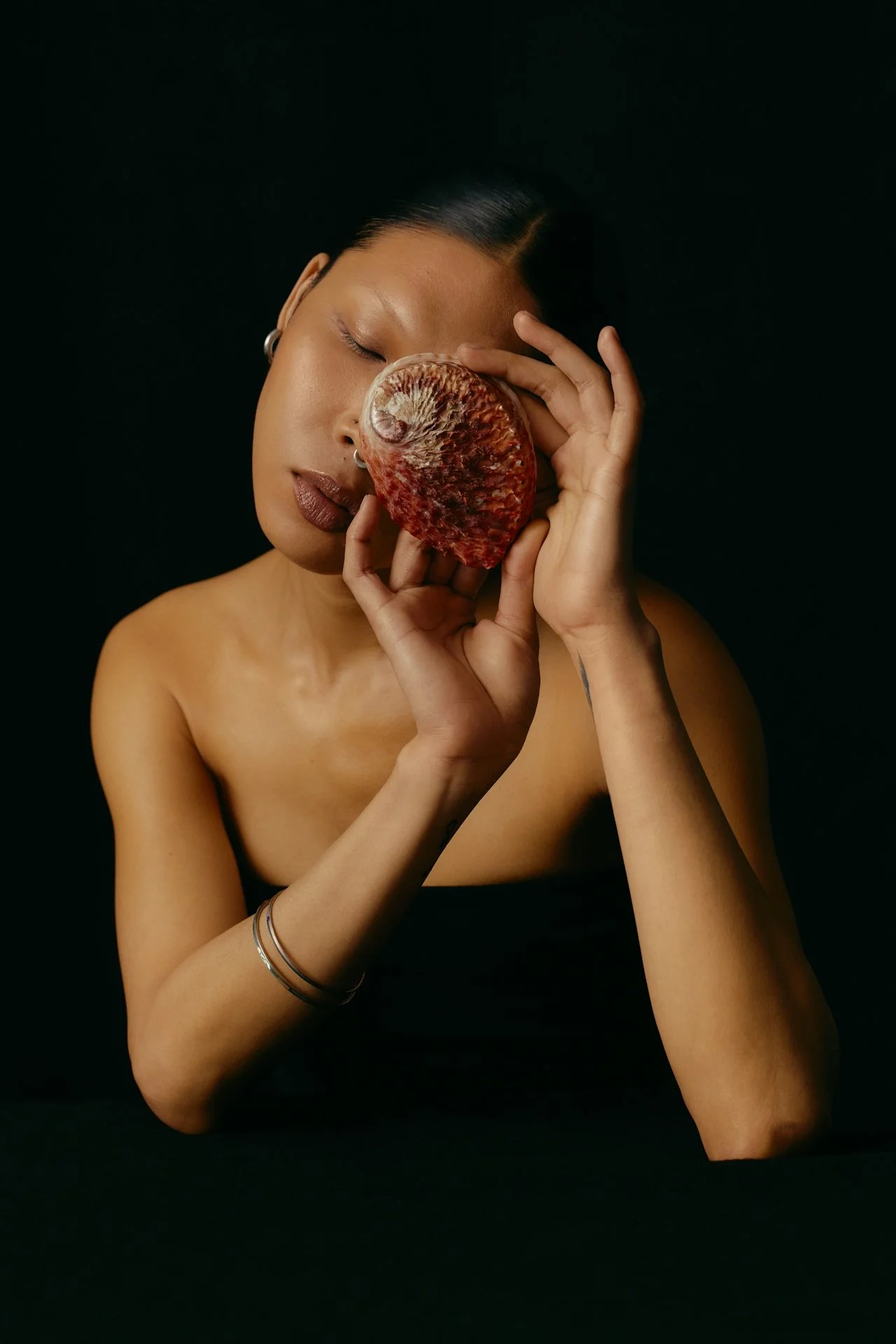 A woman with dark hair and brown skin, wearing hoop earrings and bracelets, holds a large conch shell close to her face with her eyes closed against a black background.