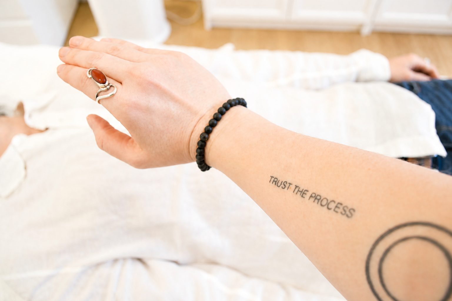 A person's arm with a tattoo reading 'TRUST THE PROCESS' and a black beaded bracelet, holding another person's hand with a silver ring featuring a large orange stone, in a cozy room.