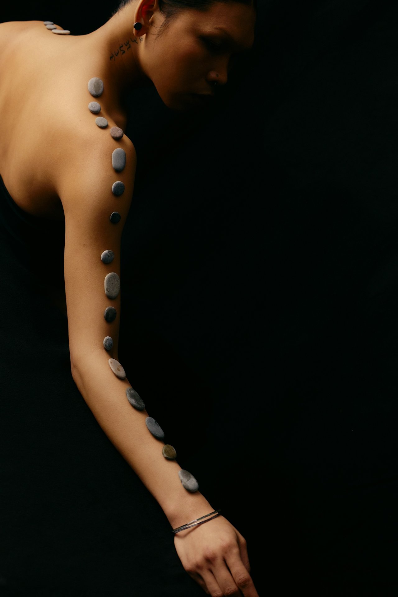 A woman lying down with her eyes closed, decorated with smooth stones aligned along her shoulder, arm, and wrist, against a black background.