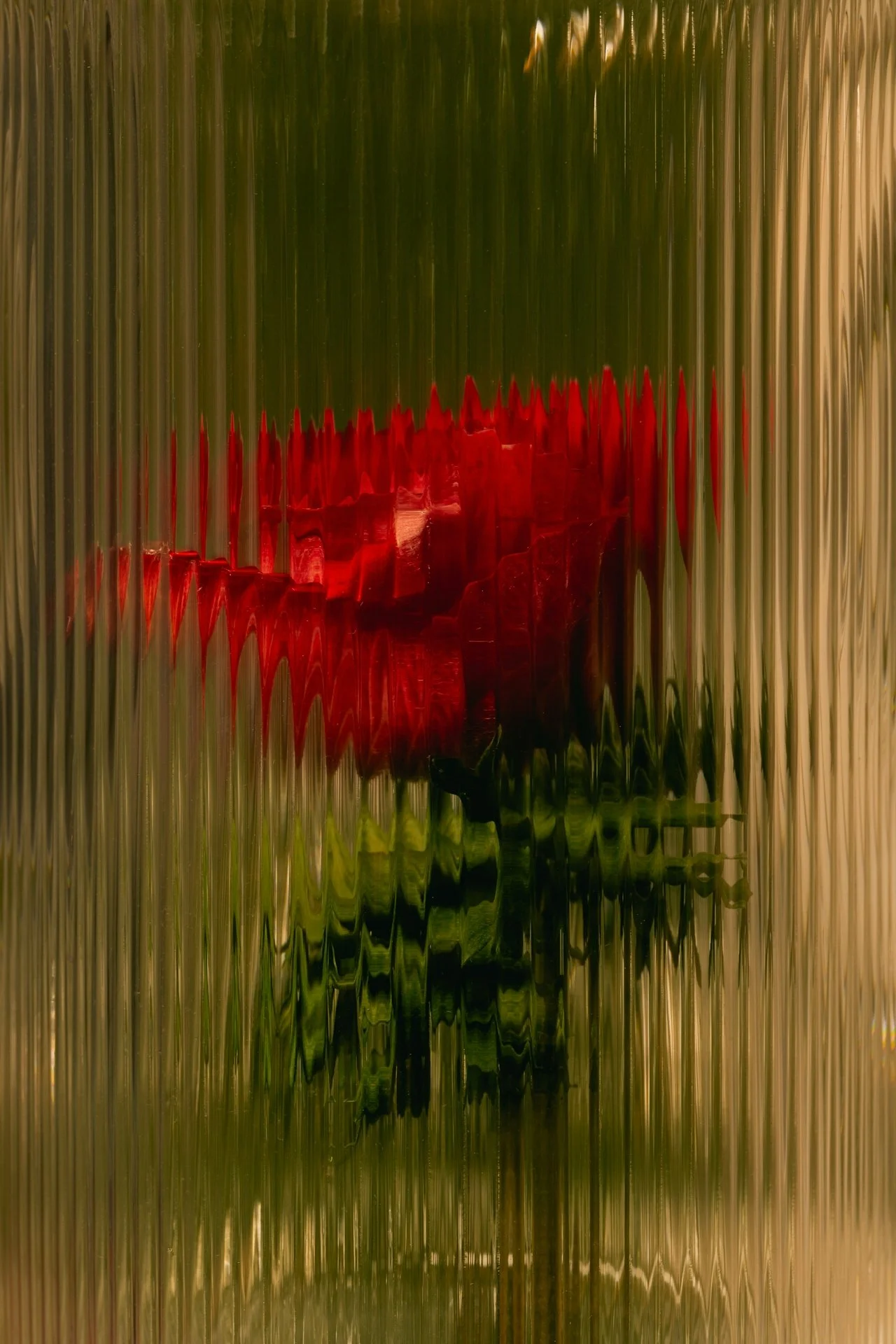 A red flower with green leaves seen through a textured, distorted glass or plastic surface.