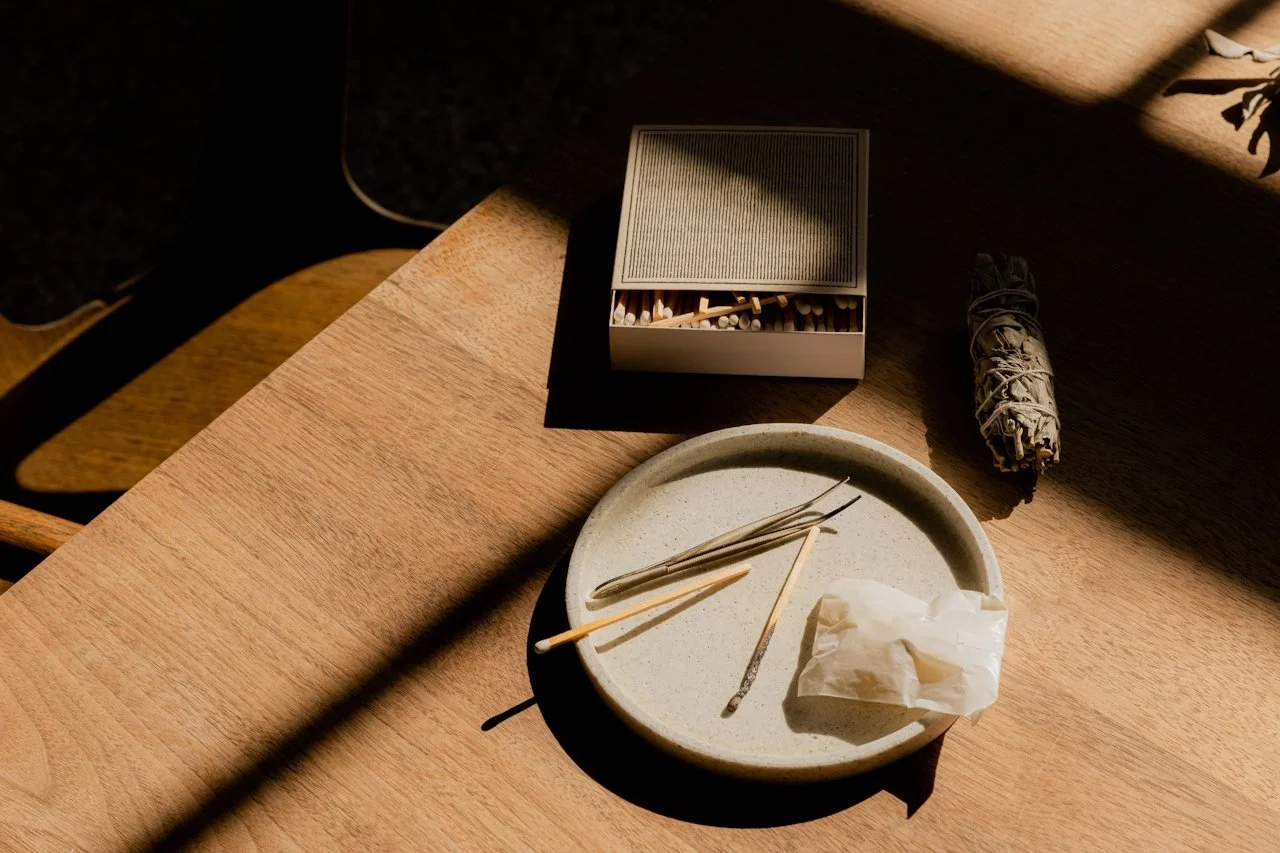 A wooden table with a round plate, candles, tissue, and an abalone shell. A box of matches and a rolled-up bundle also on the table.