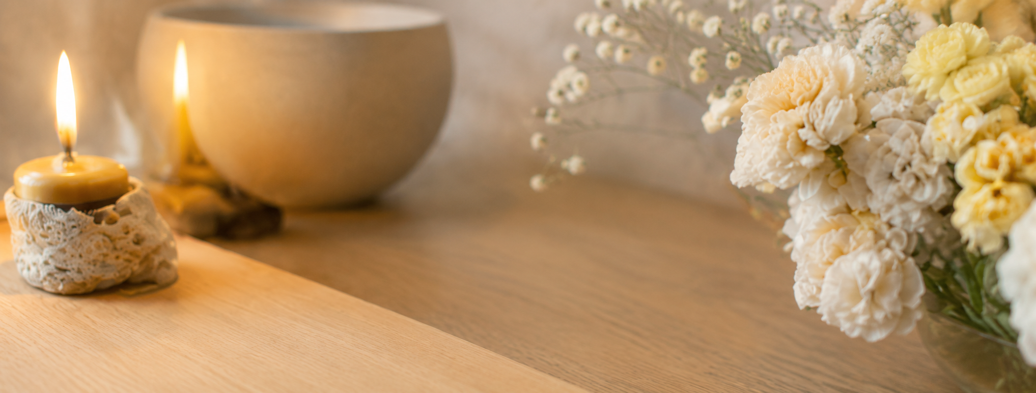 A lit candle on a ceramic holder with lace pattern, next to a white vase and a bouquet of white and yellow flowers on a wooden surface.
