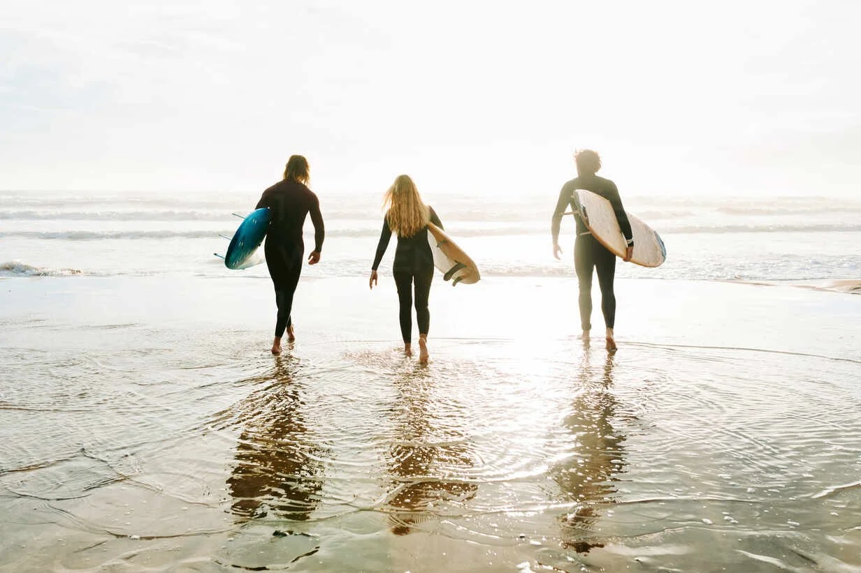 Three people are walking out of the ocean at sunset, each carrying a surfboard.