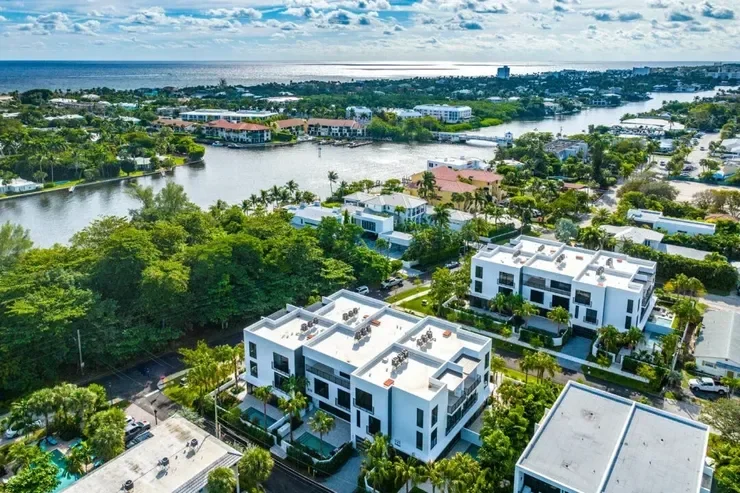 Bird's eye view of modern white apartment buildings surrounded by lush greenery with a river in the background under a partly cloudy sky.