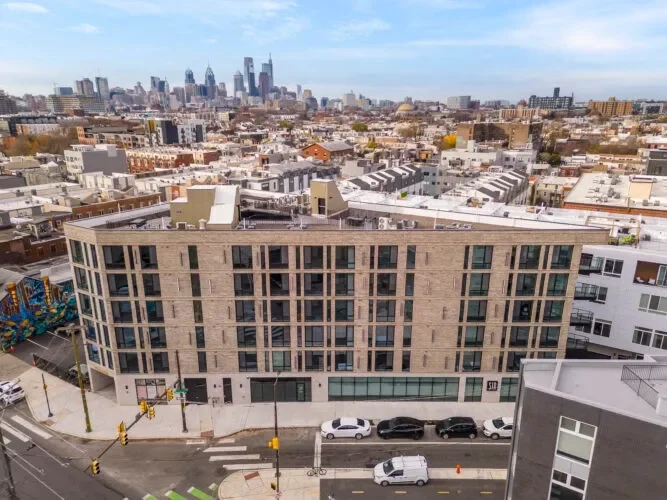 City skyline with tall skyscrapers in the distance, and a beige modern apartment building with many windows in the foreground, located on a street corner with parked cars.