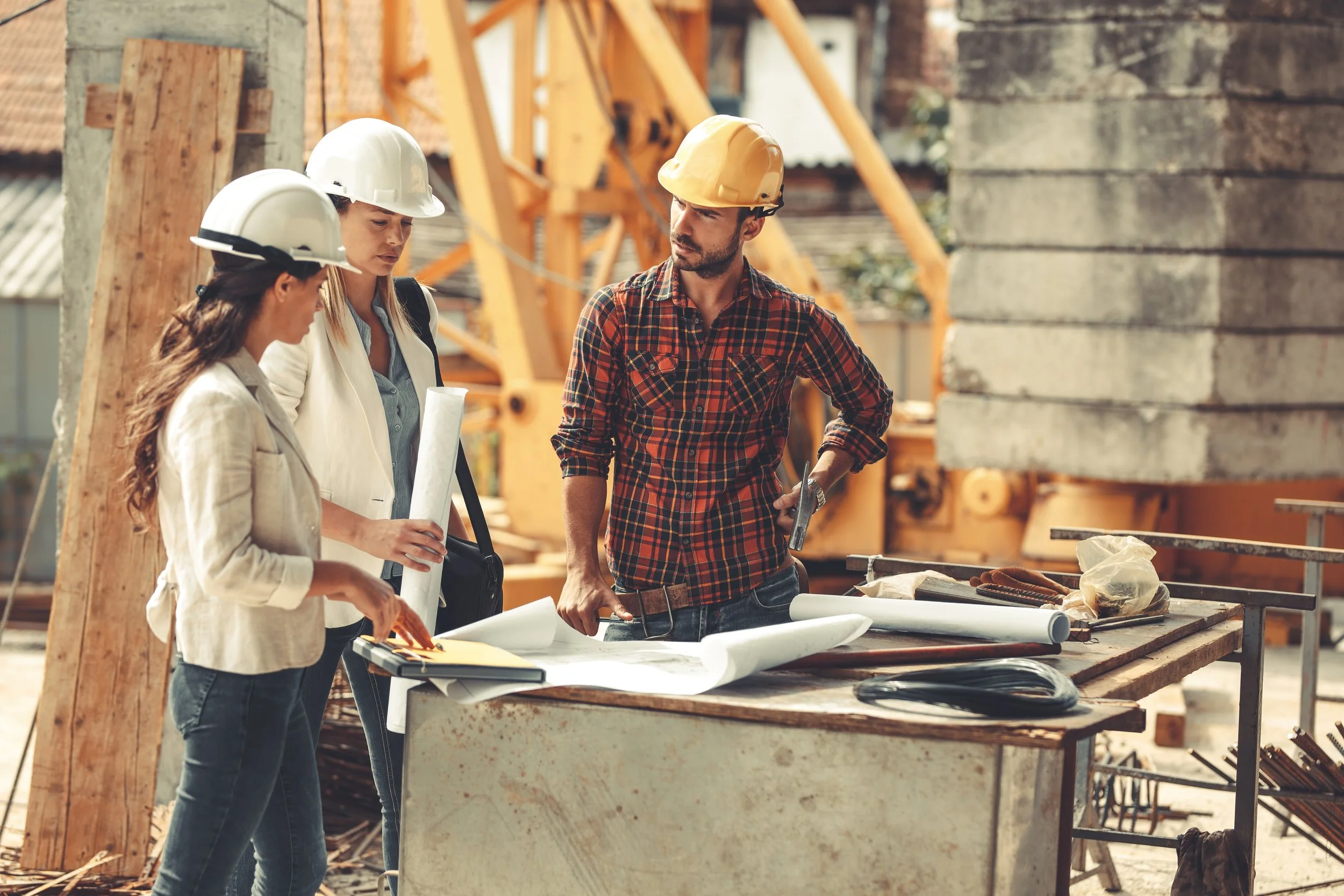 Three construction workers, two women and one man, wearing hard hats and discussing plans at a construction site with wooden and metal structures around.