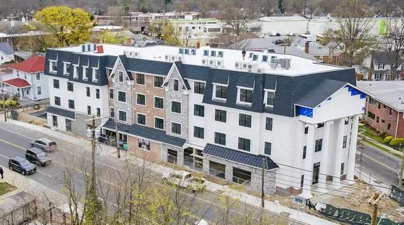 A large multi-story residential building under construction in an urban area, with several parked cars on the street and construction materials nearby.