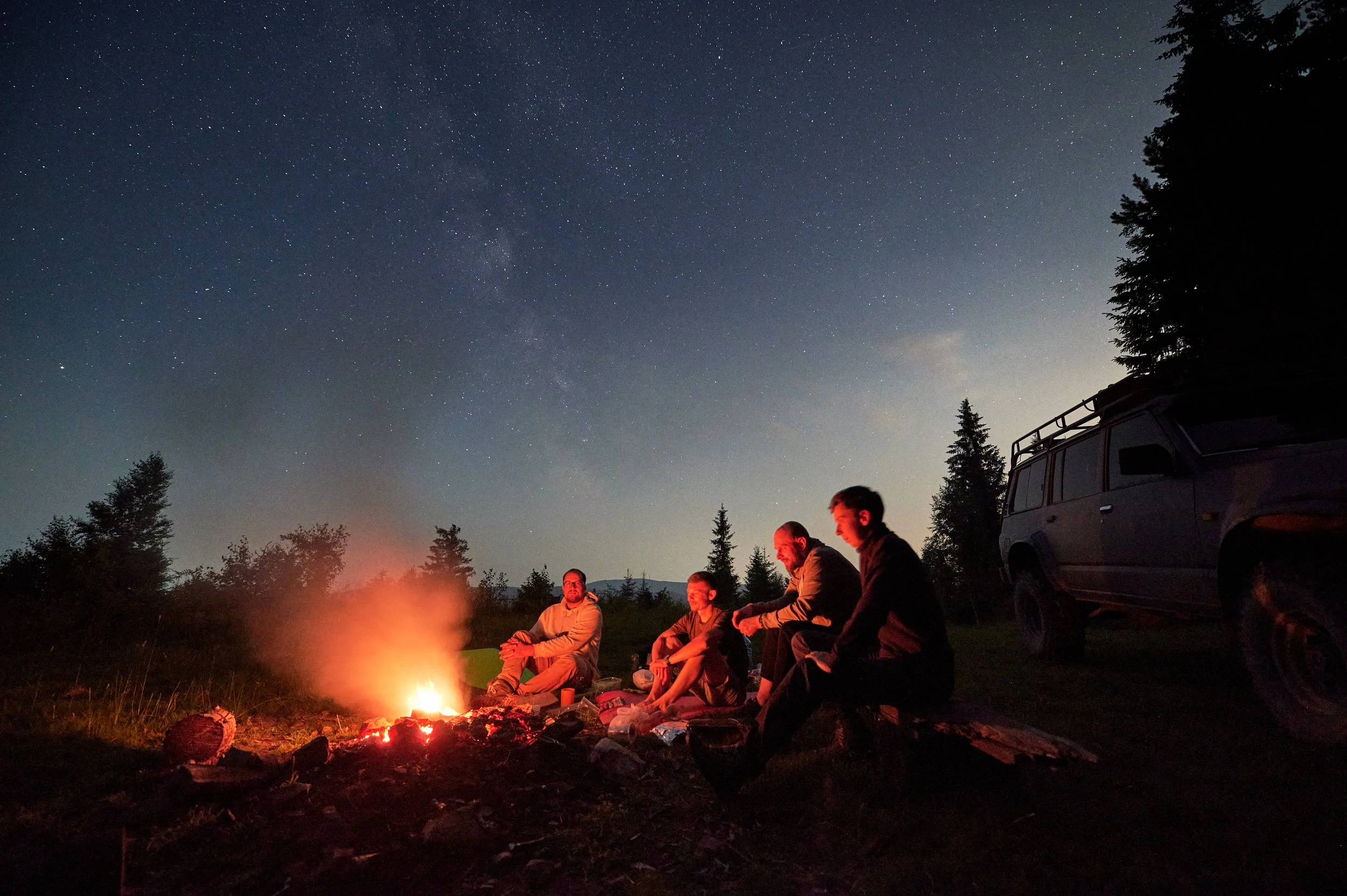 Group of four people sitting around a campfire at night in a forested area with trees and a starry sky overhead.
