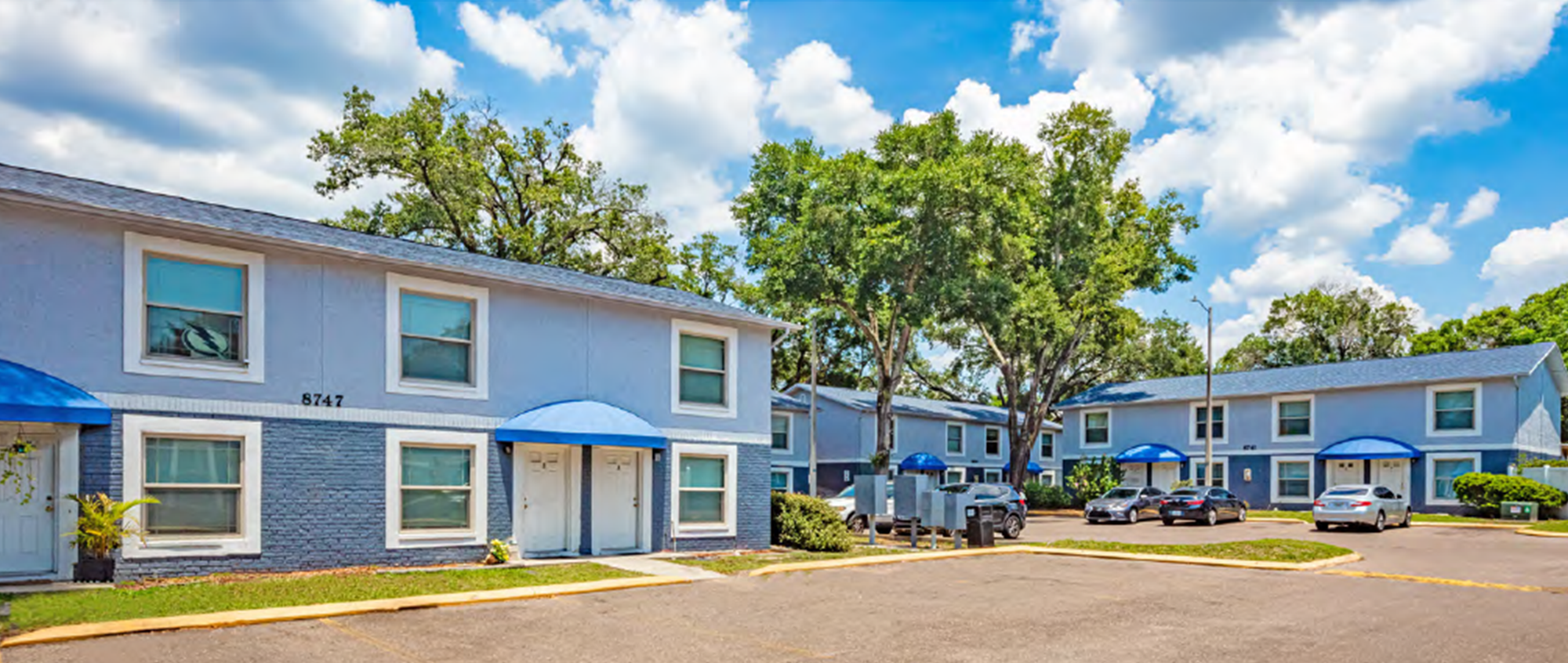Apartment complex with blue buildings, parking lot with cars, large trees, and a partly cloudy sky.