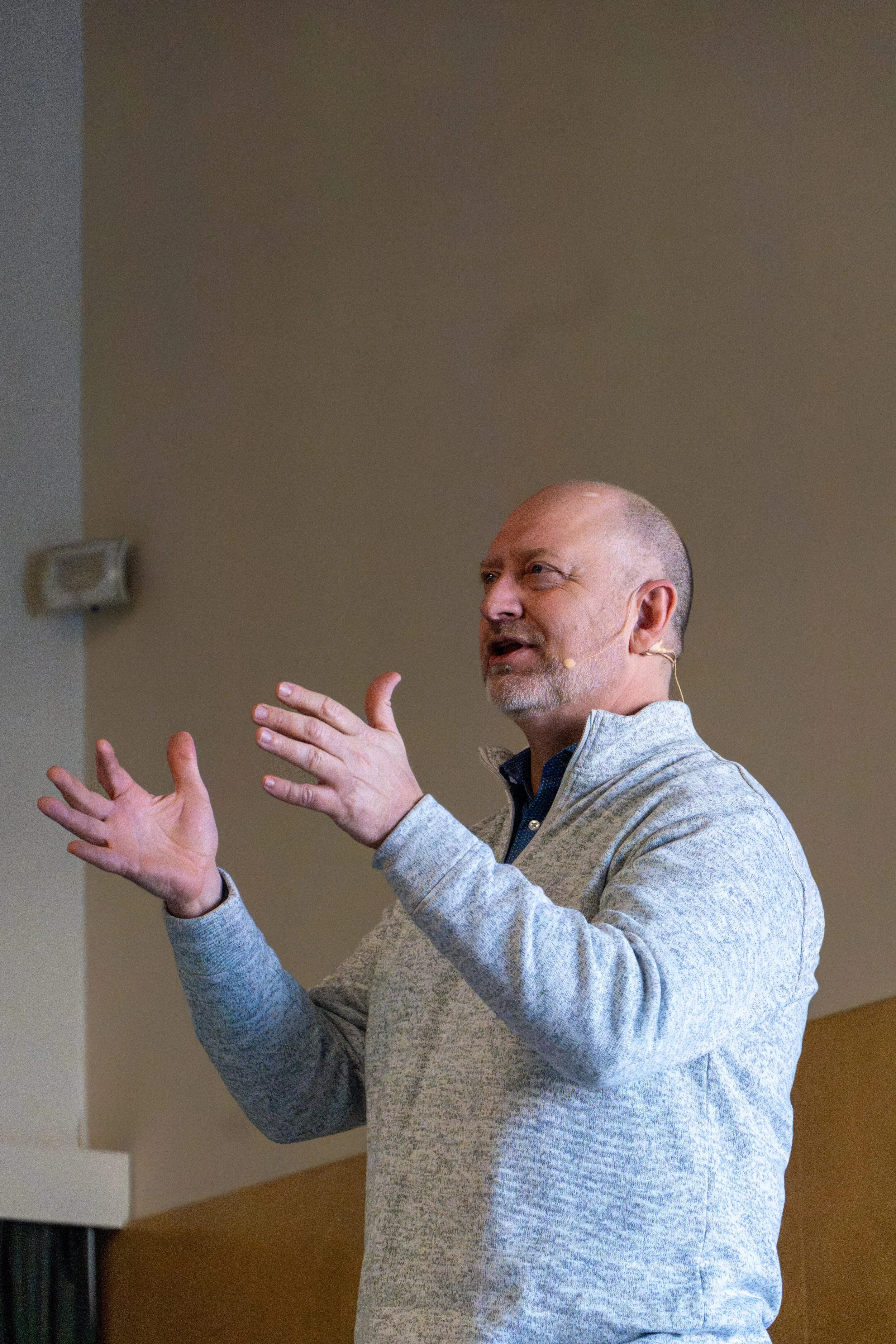 A man with a shaved head and gray beard speaking and gesturing with his hands during a presentation or talk, wearing a light gray sweater and headset microphone in a room with beige walls.