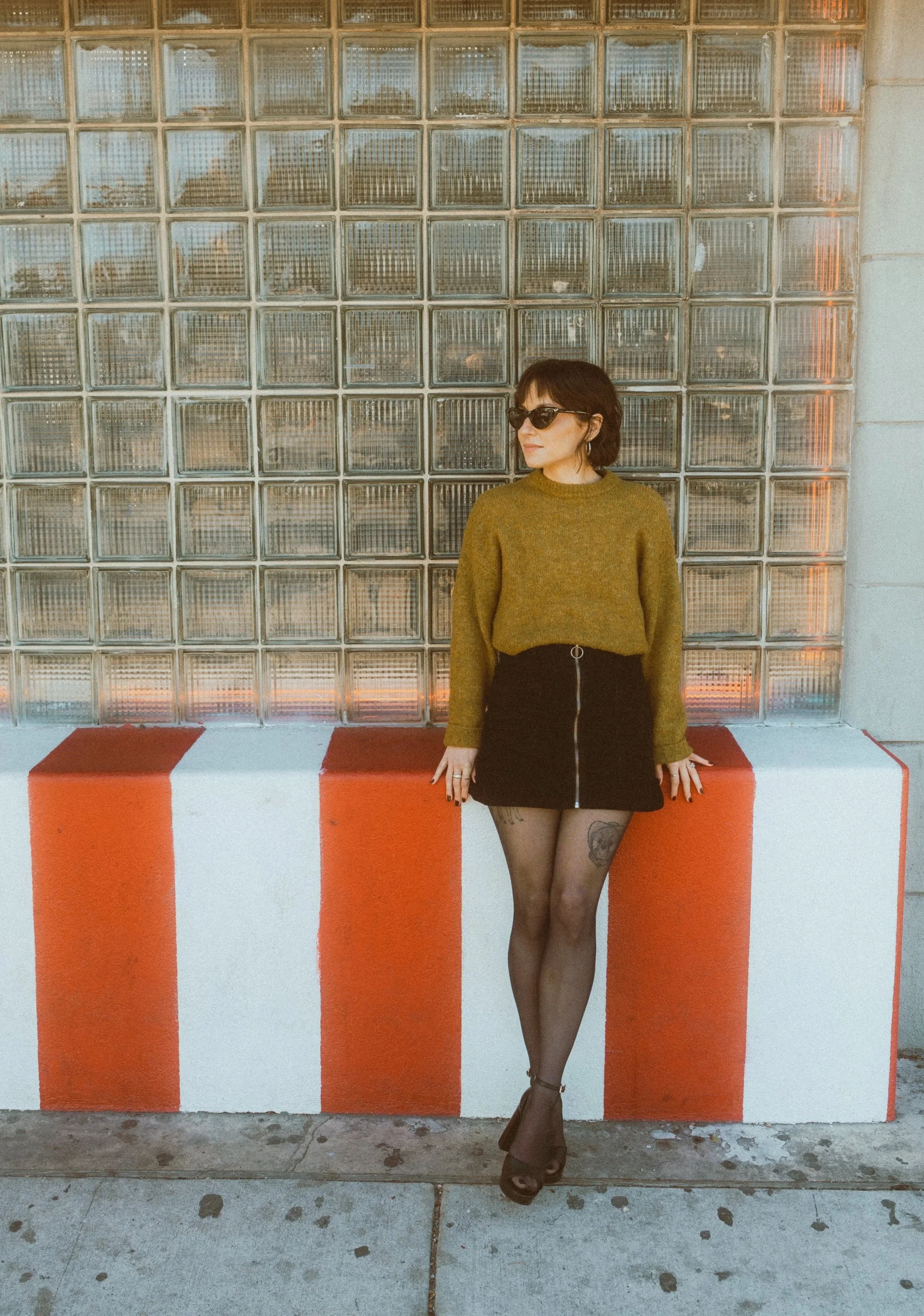 A person standing against a wall with a glass block window, wearing sunglasses, a green sweater, a black skirt with a zipper, tights, and high heels.