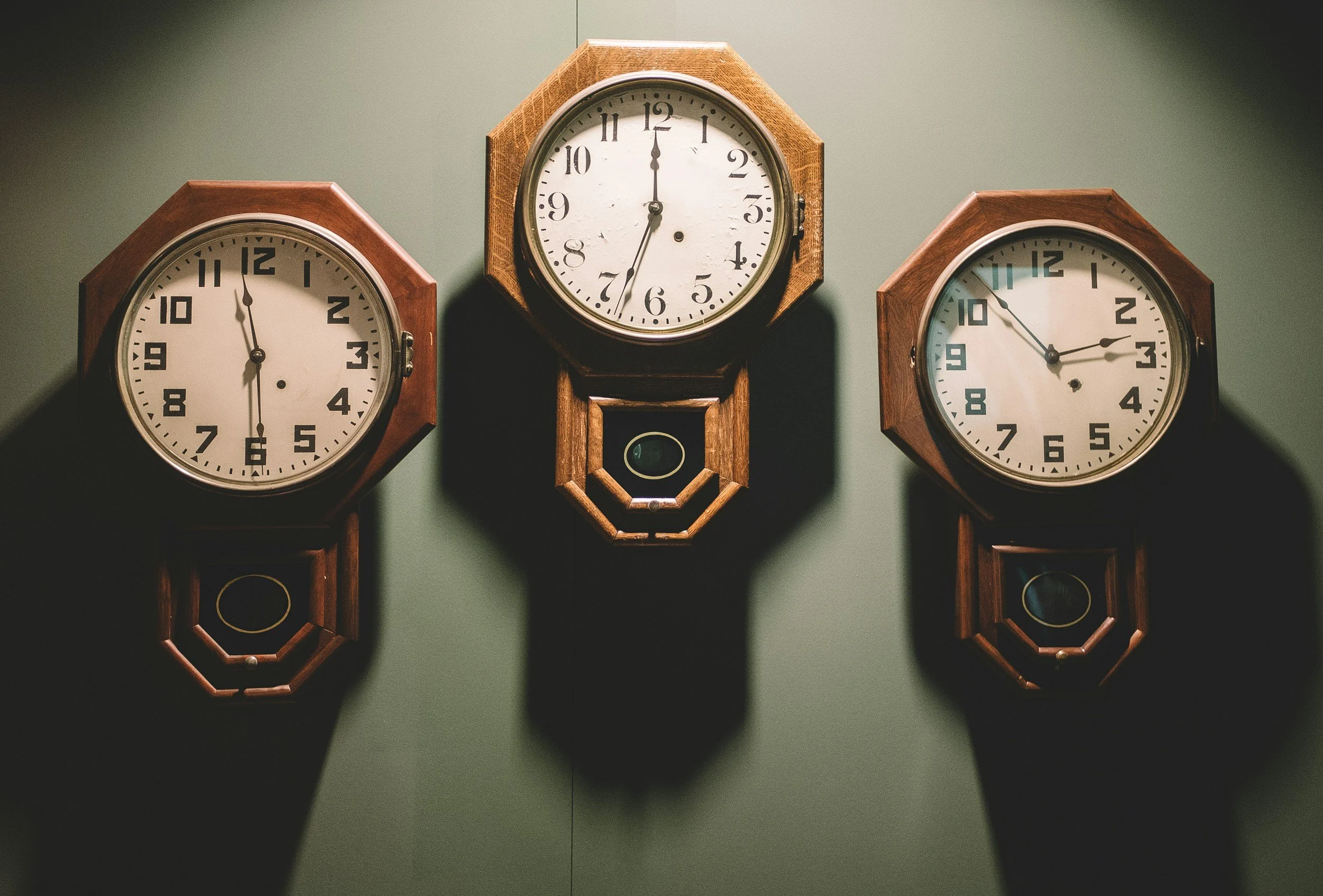 Three vintage wooden wall clocks mounted on a green wall, casting shadows.