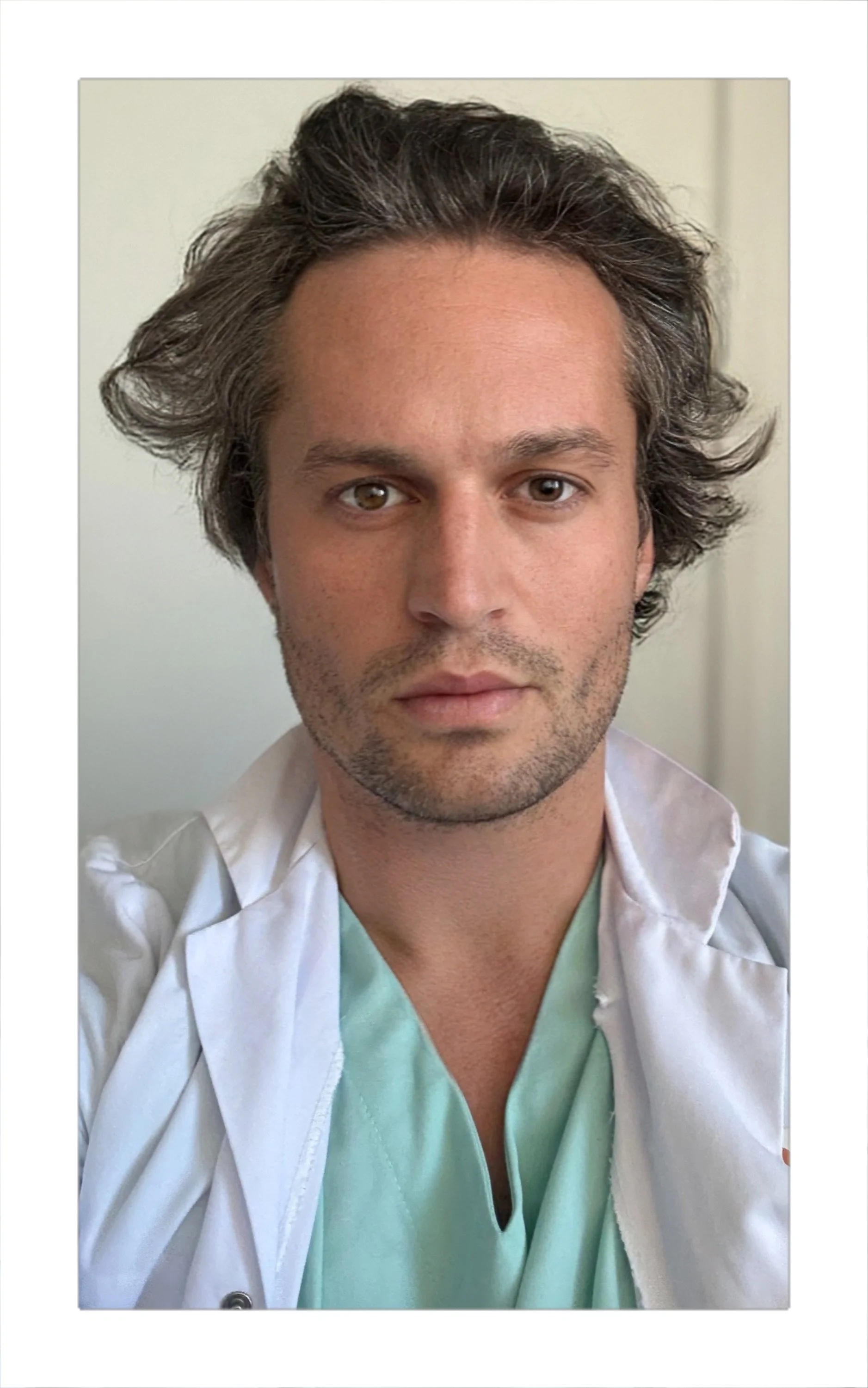 A man with wavy dark hair, a slight beard, and light brown eyes wearing medical scrubs and a white lab coat.